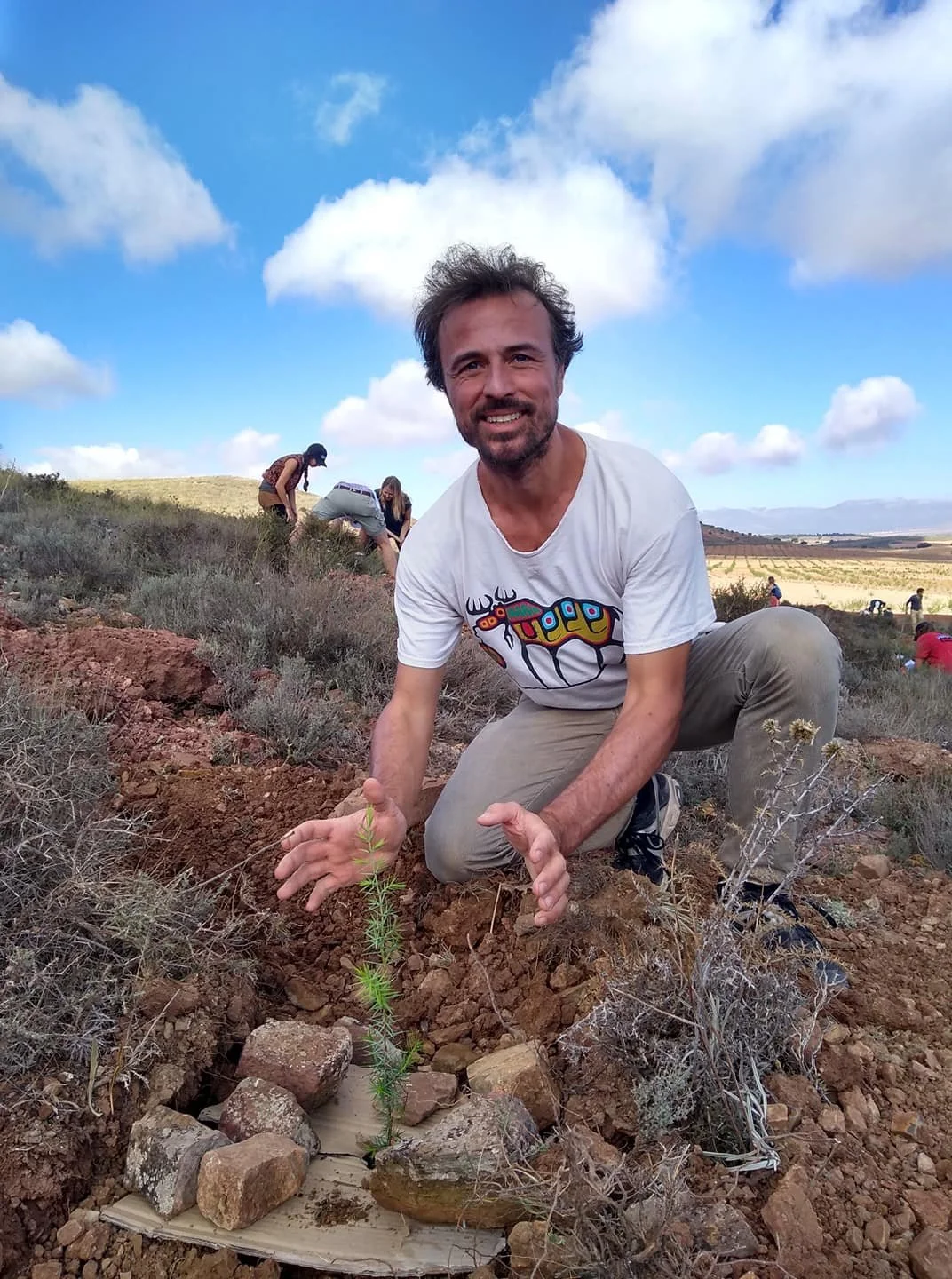 A man kneeling on the ground and planting a small seedling in a dry, rocky landscape under a partly cloudy sky, with other people working in the background.