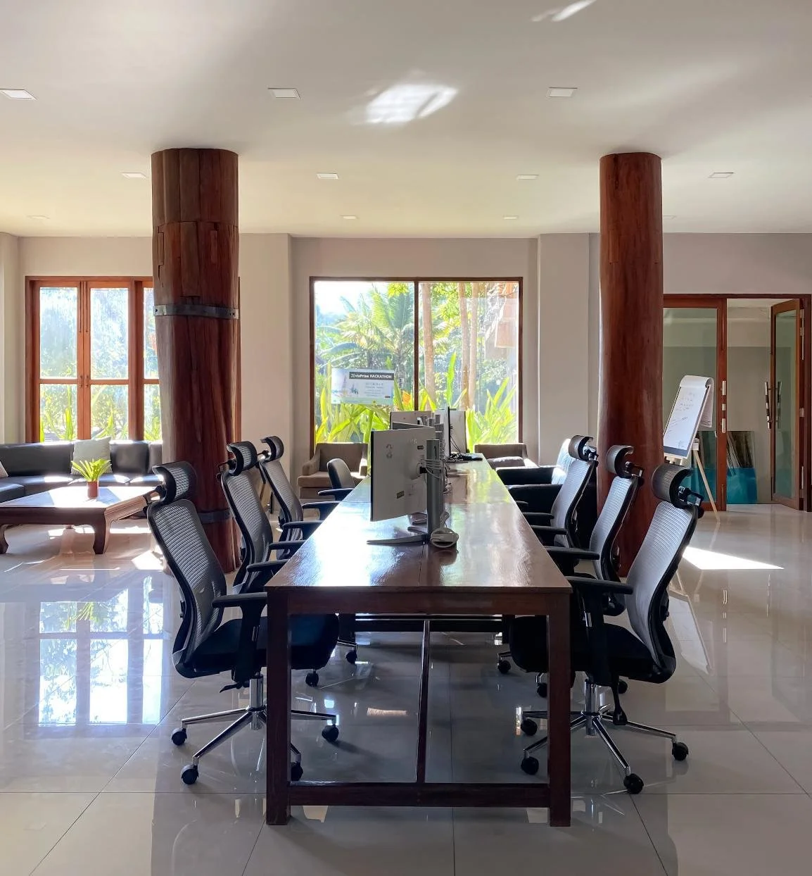 Modern conference room with large wooden table, office chairs, computer monitors, and large windows showing outdoor greenery.