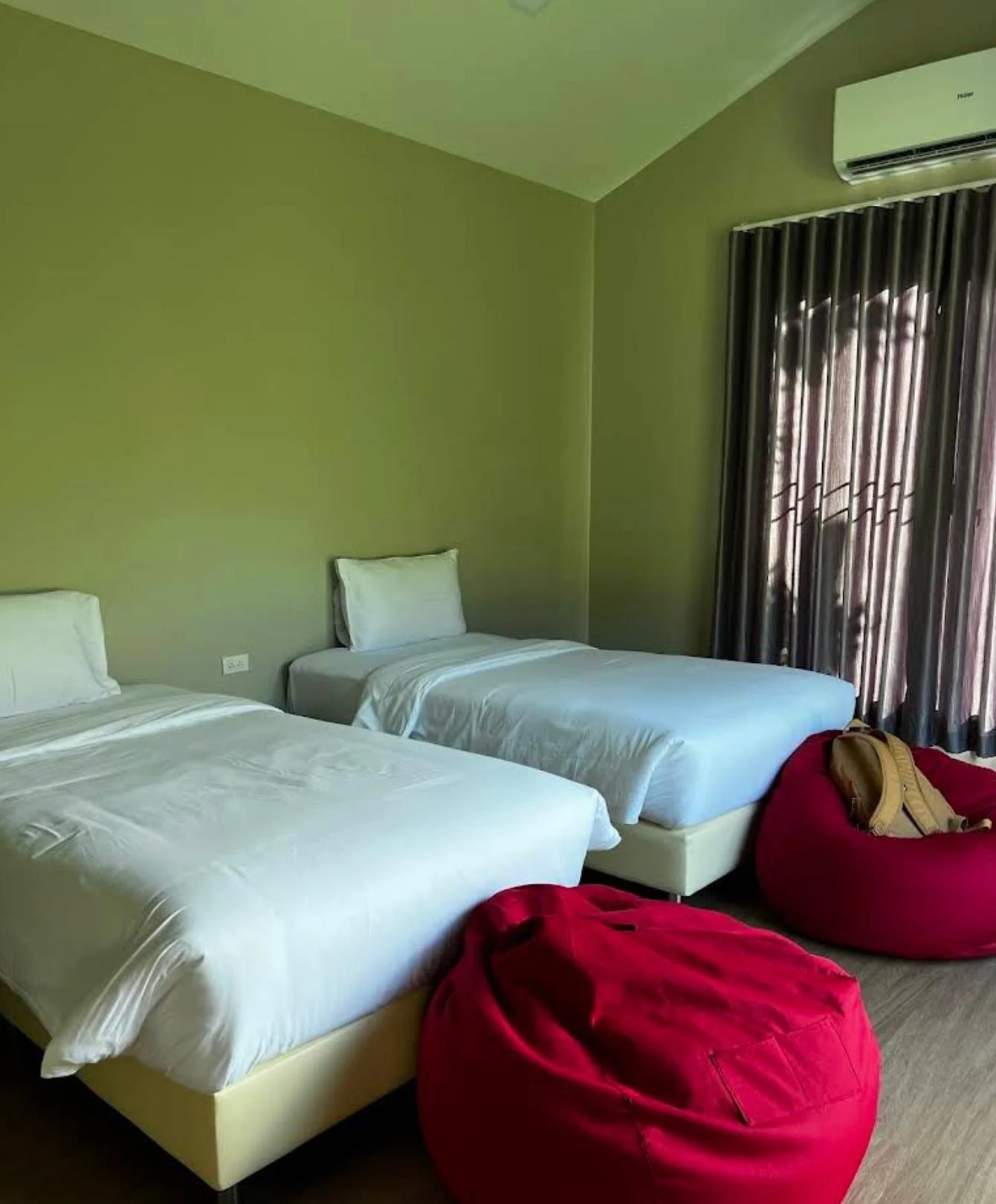 Two single beds with white bedding and pillows in a hotel room, with a large red bean bag and a maroon bean bag chair with a backpack on top, near a window with dark curtains and an air conditioner above.