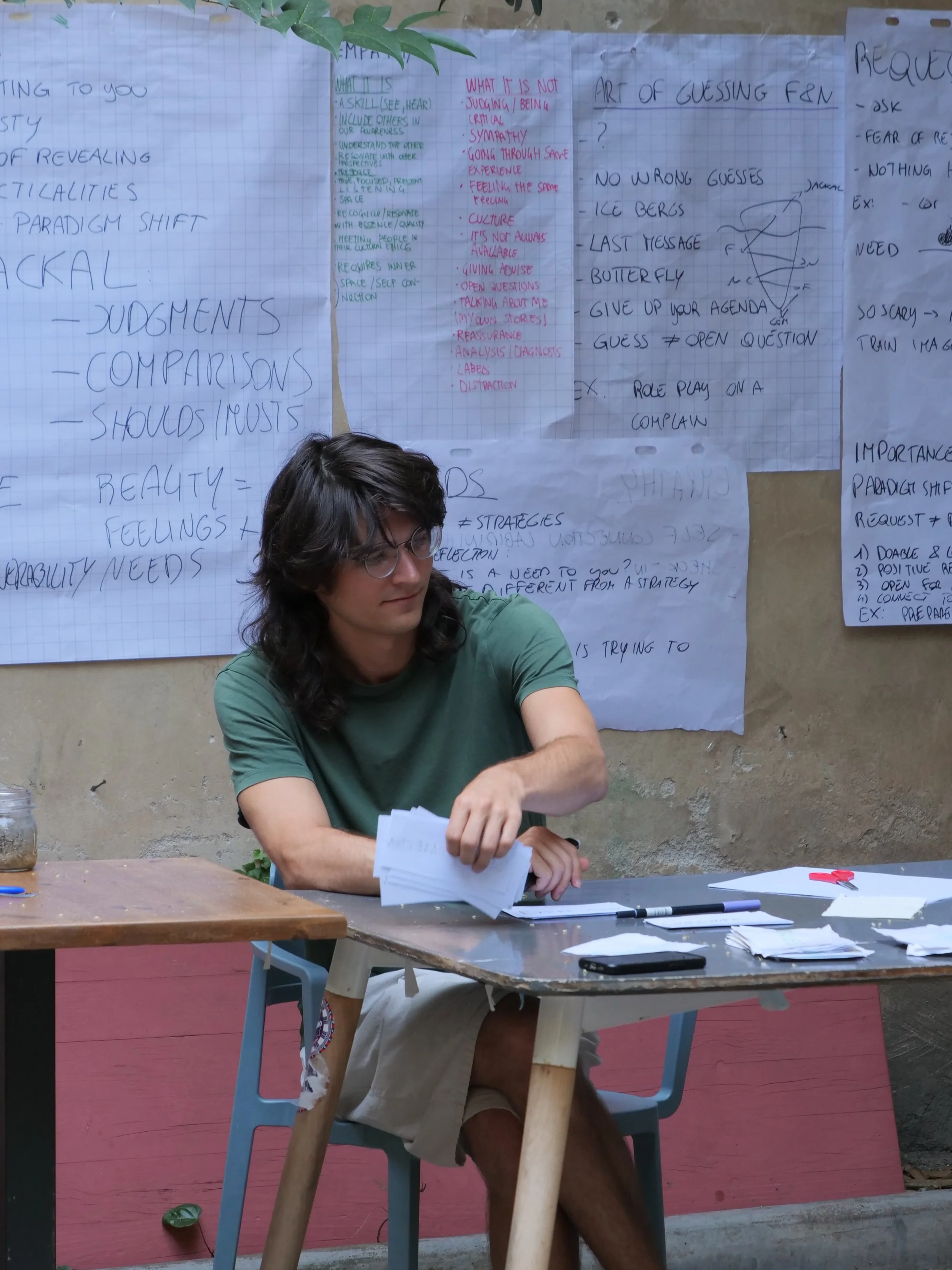 A young man with shoulder-length dark hair and glasses sits at a desk with papers, a pen, and a phone. Behind him are sheets of paper with handwritten notes and diagrams taped to the wall.