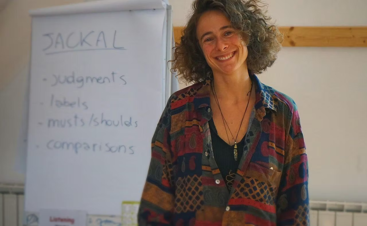 A woman with curly hair smiling in front of a whiteboard with the word 'JACKAL' and bullet points on judgments, labels, musts/shoulds, and comparisons written on it.