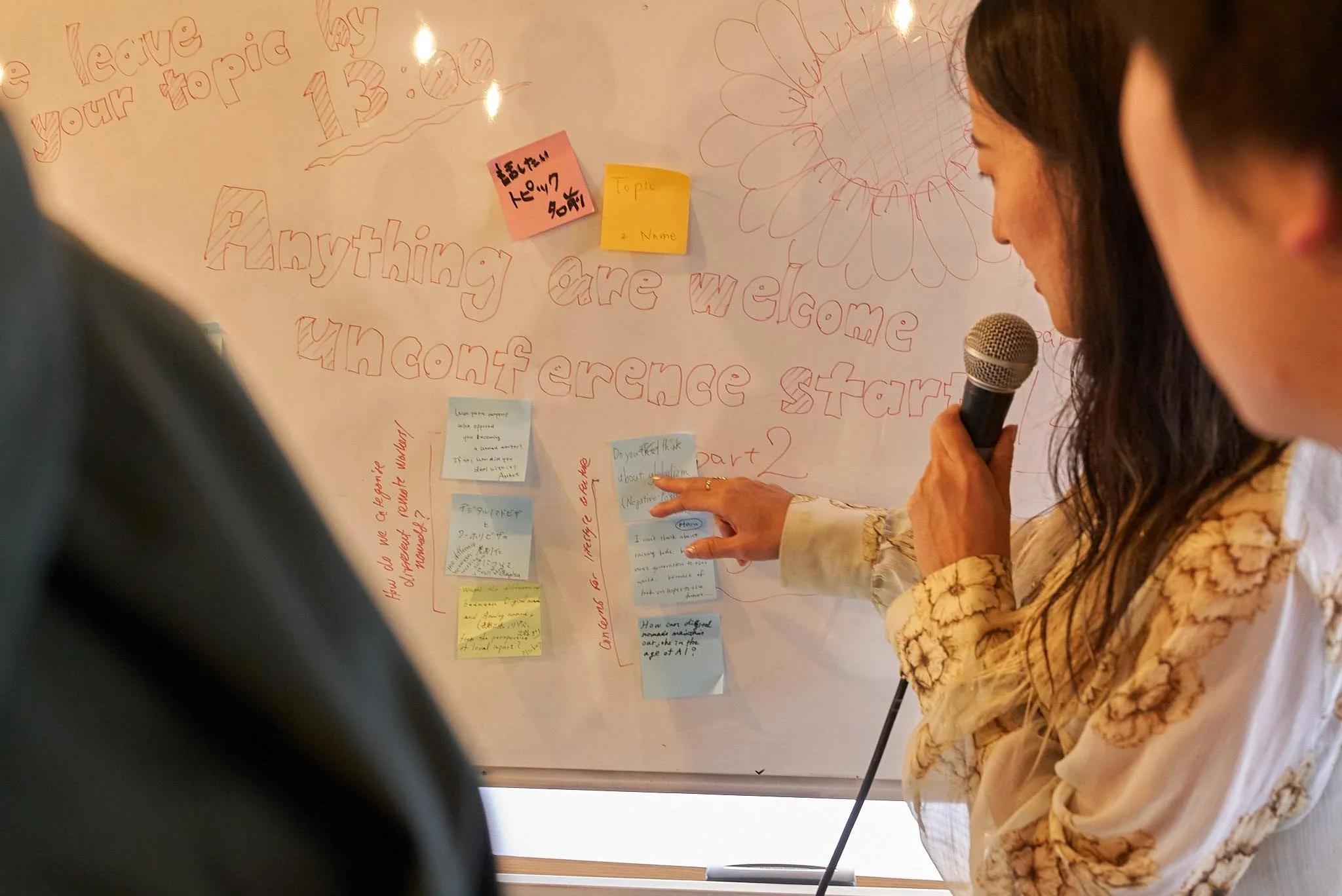 A woman holding a microphone and pointing to a list of sticky notes on a large paper titled 'Anything are welcome unconference start' with additional handwritten notes and drawings.
