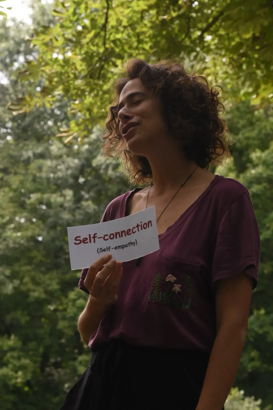 A woman with curly brown hair holding a sign that reads "Self-connection (Self-empathy)" while standing outdoors among green trees.