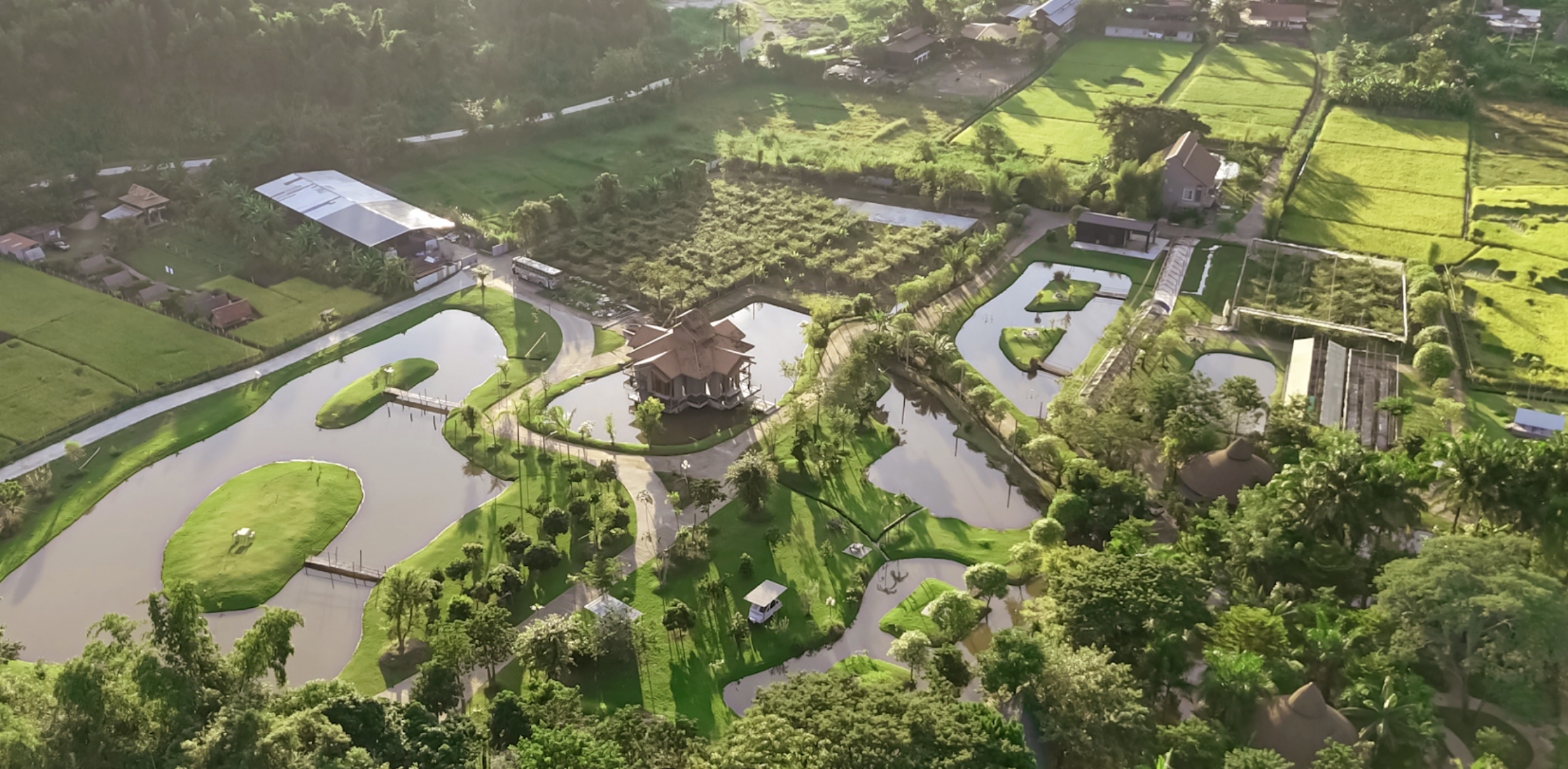 Aerial view of a lush resort with interconnected ponds, a central pavilion, walking paths, surrounded by trees, fields, and small buildings in the countryside.