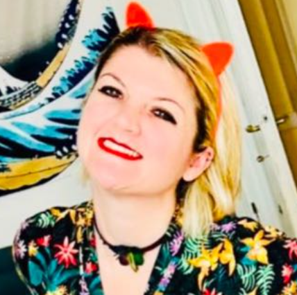 Woman with blonde hair smiling, wearing red devil horn headband and colorful floral top, in a room with a mirror and curtains.