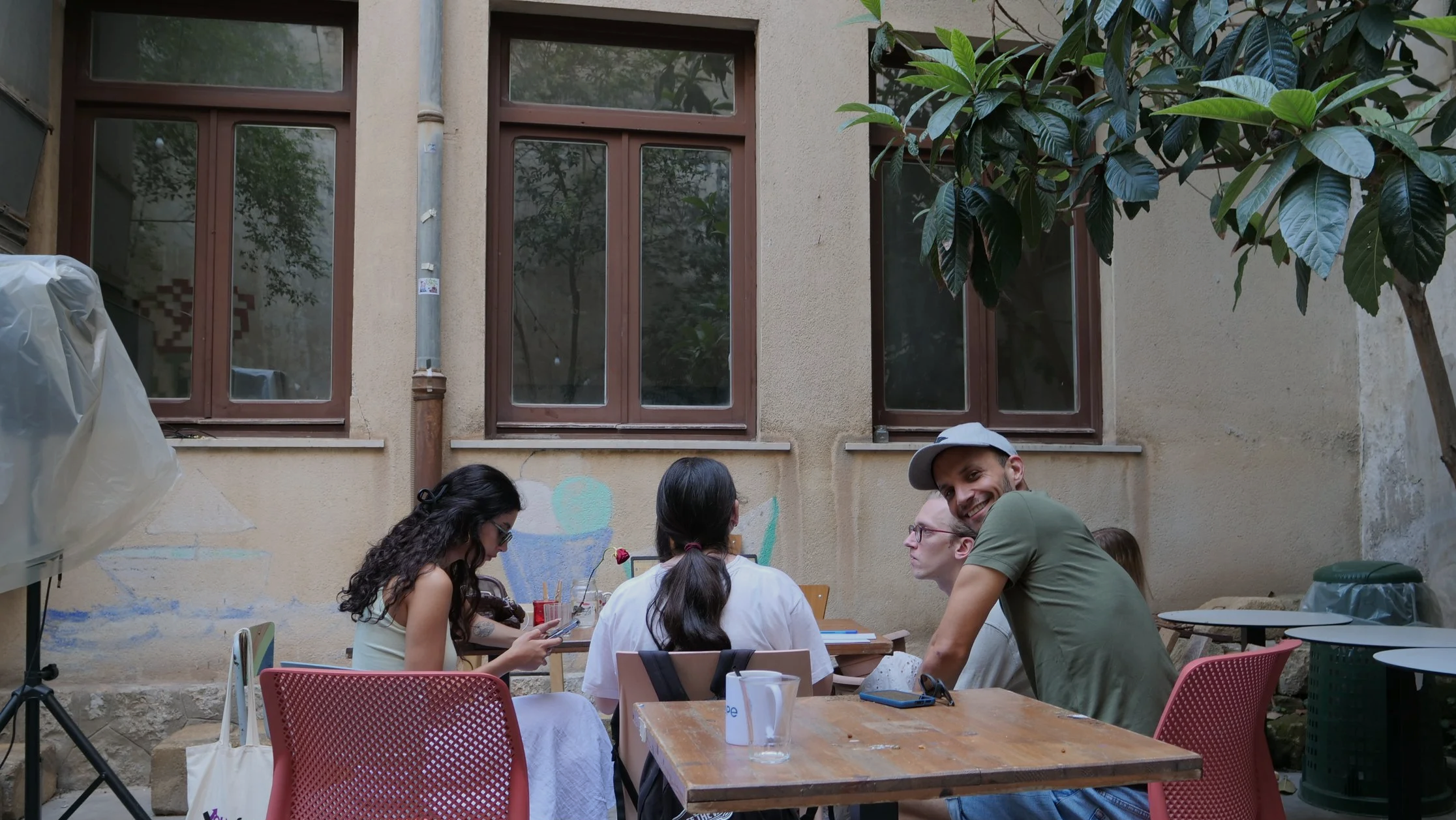 People sitting and socializing at outdoors patio table with chairs, beer mugs, and drinks, in front of a beige wall with four windows and lush green tree.