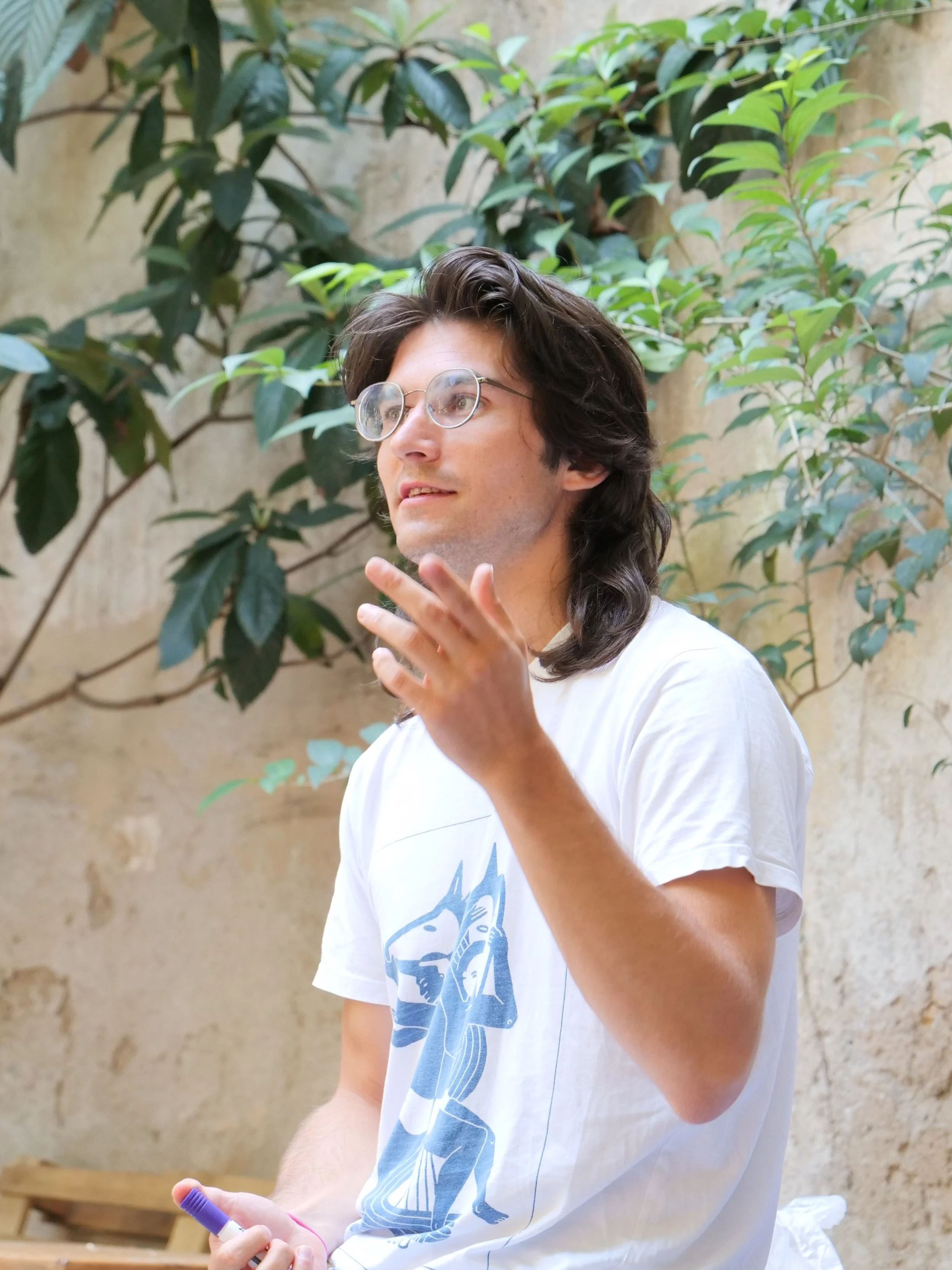 A young man with long dark hair and glasses, wearing a white T-shirt with a graphic design, is gesturing with his right hand while holding a marker in his left hand. He is seated outdoors against a backdrop of green leafy plants and a textured wall.