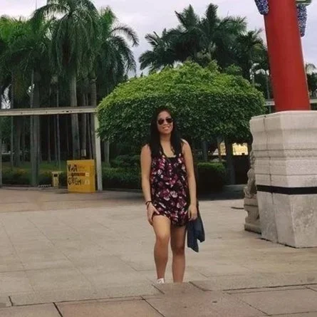 A woman in a floral dress and sunglasses standing outdoors near a stone structure and greenery.