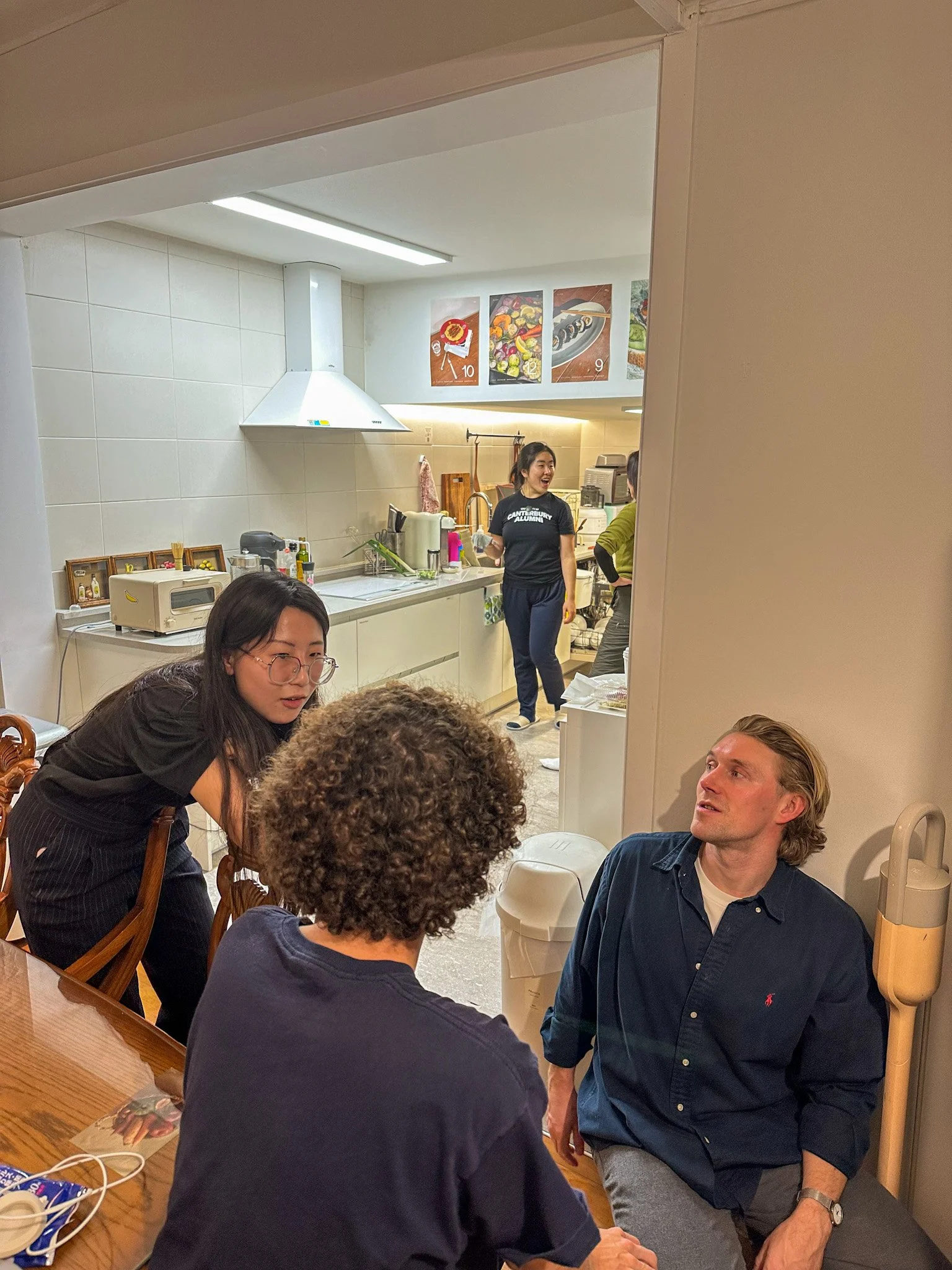Four people engaging in a conversation in a dining area, with a kitchen visible in the background.