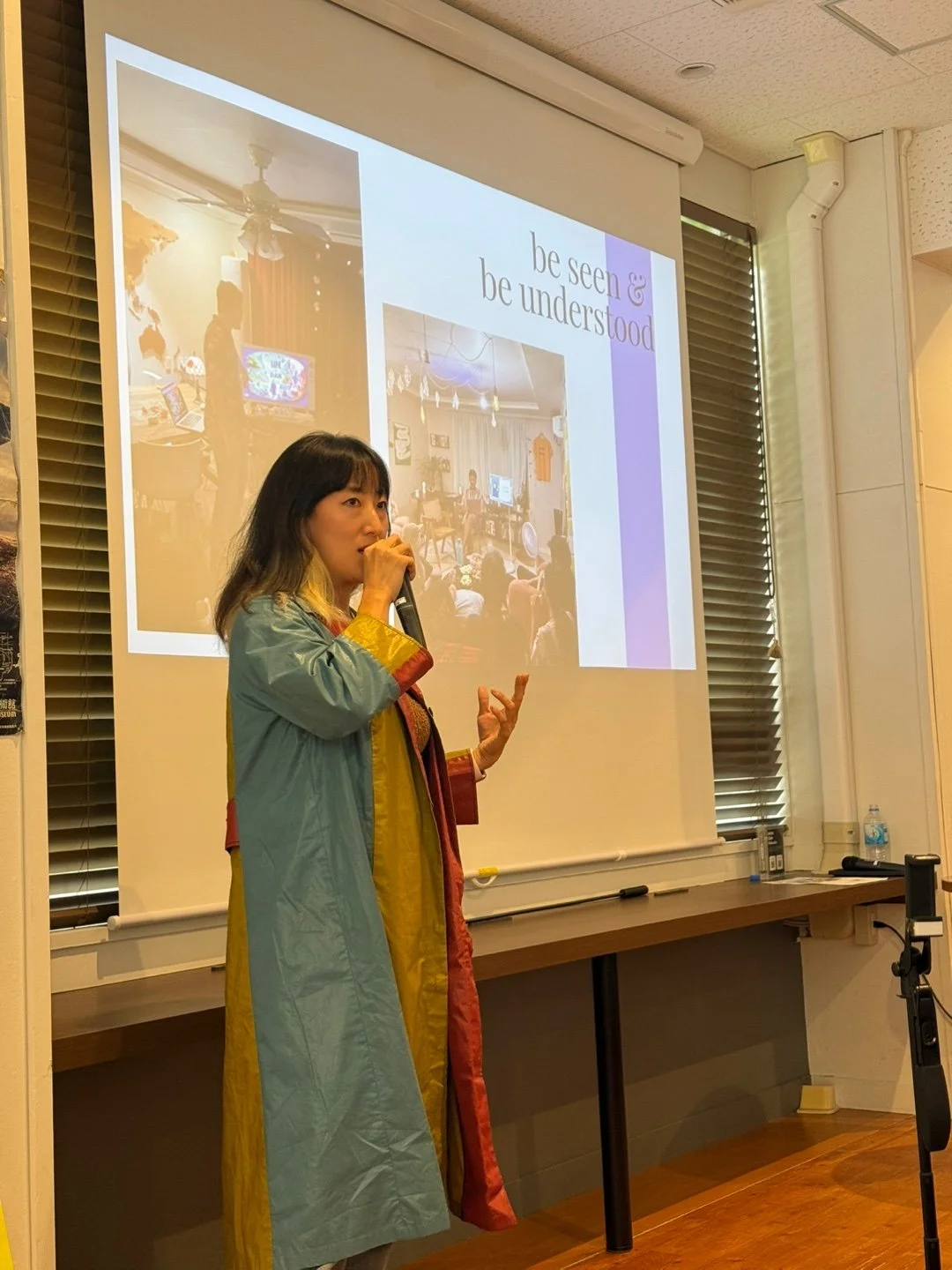 A woman giving a presentation in front of a large screen with slides. She is holding a microphone and gesturing with her hand. The slide contains two photos and the text 'be seen & be understood'.