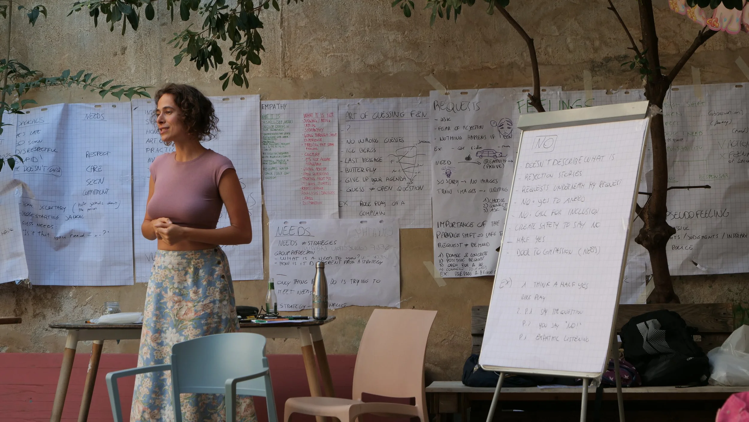 A woman in a pink shirt and floral skirt standing in front of a wall with several sheets of paper with handwritten notes attached to it, and a whiteboard with notes. There are chairs and a table with water bottles and a thermos in front of her.