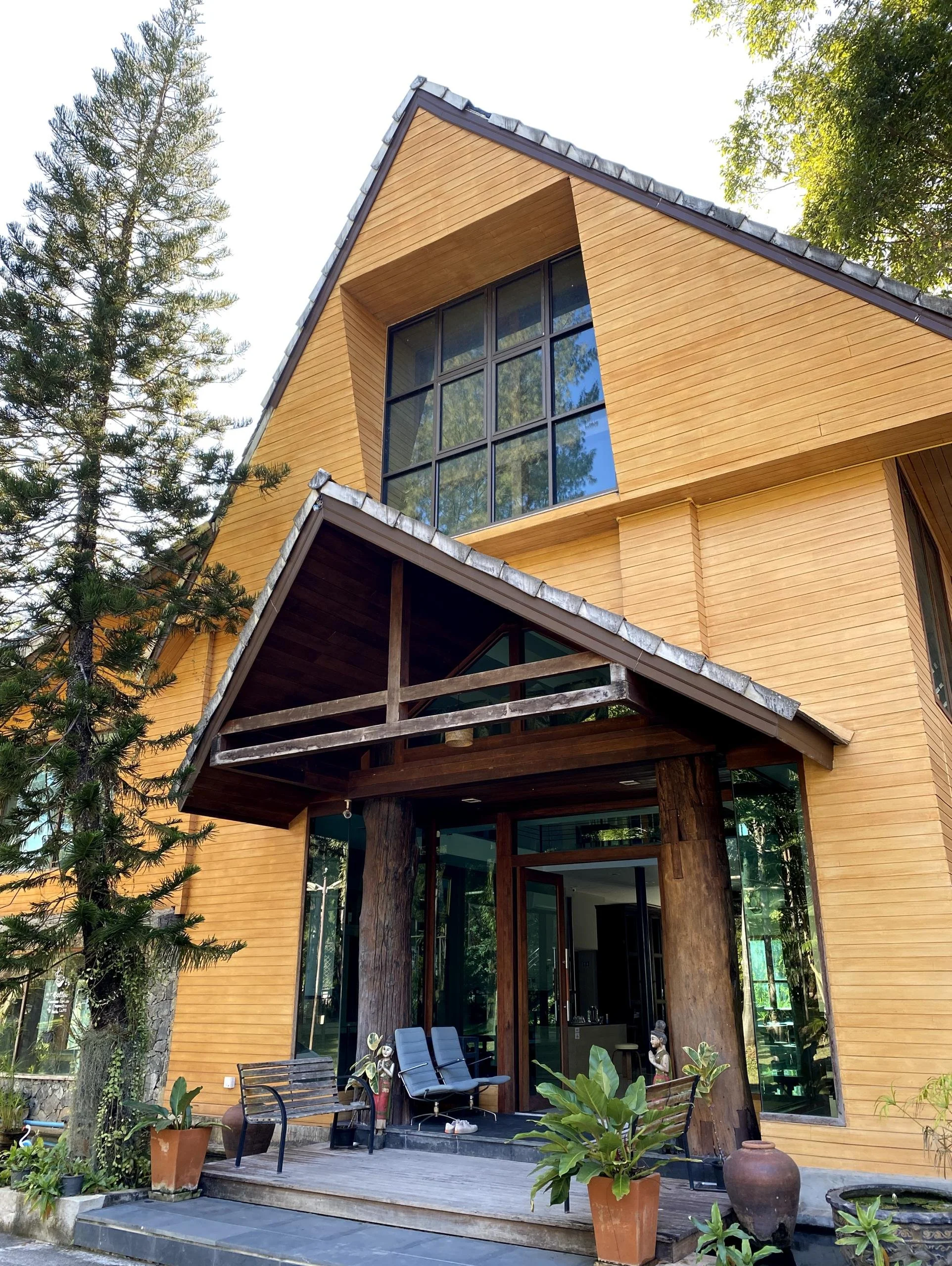 A modern wooden house with large glass windows, a sloped roof, and a small porch with seating, surrounded by greenery.