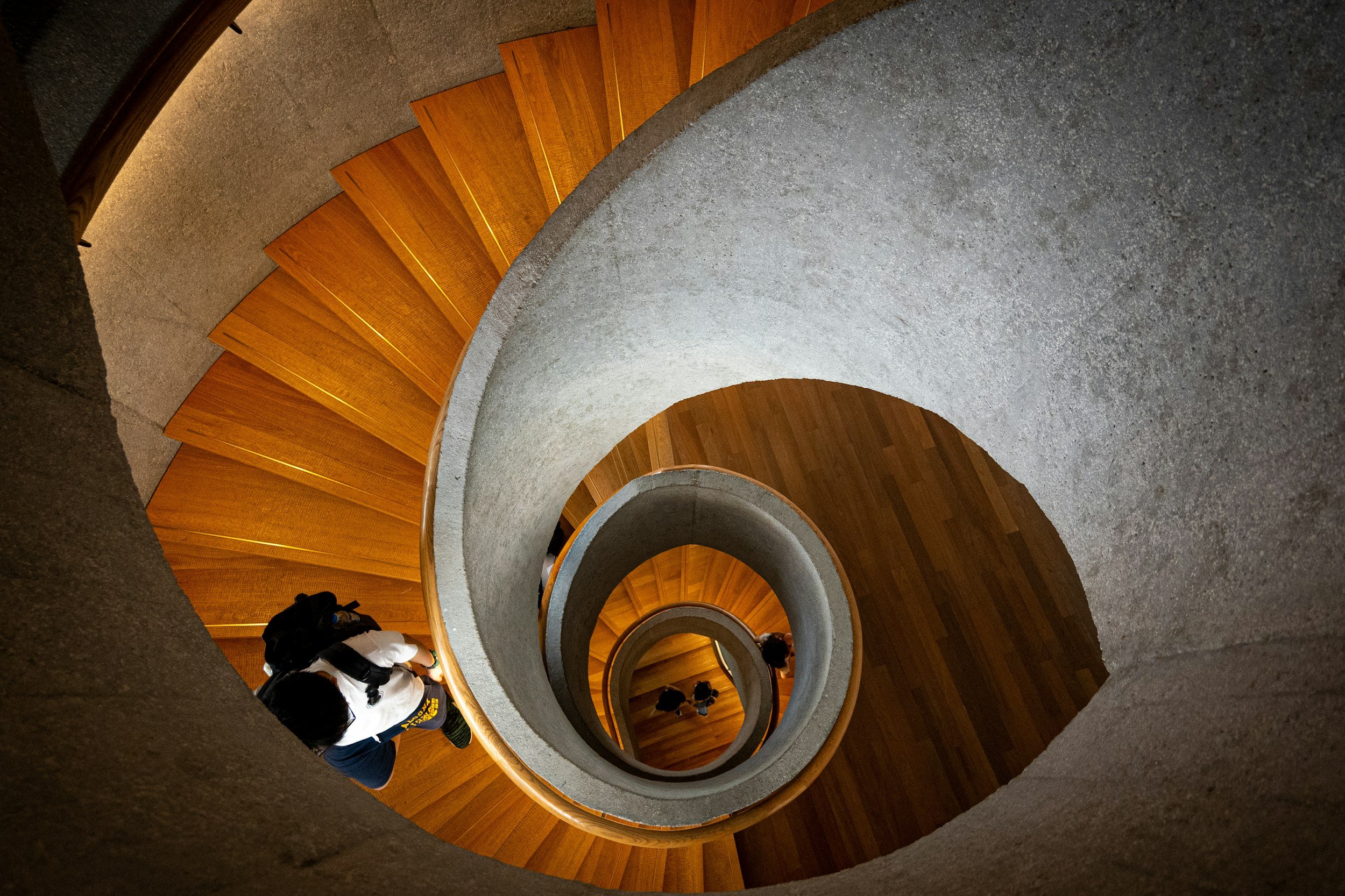 Spiral staircase with wooden steps and concrete sides, viewed from above with people walking down.