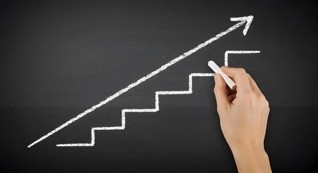 Hand drawing an upward trending arrow with chalk on blackboard, along with staircase and line graph.