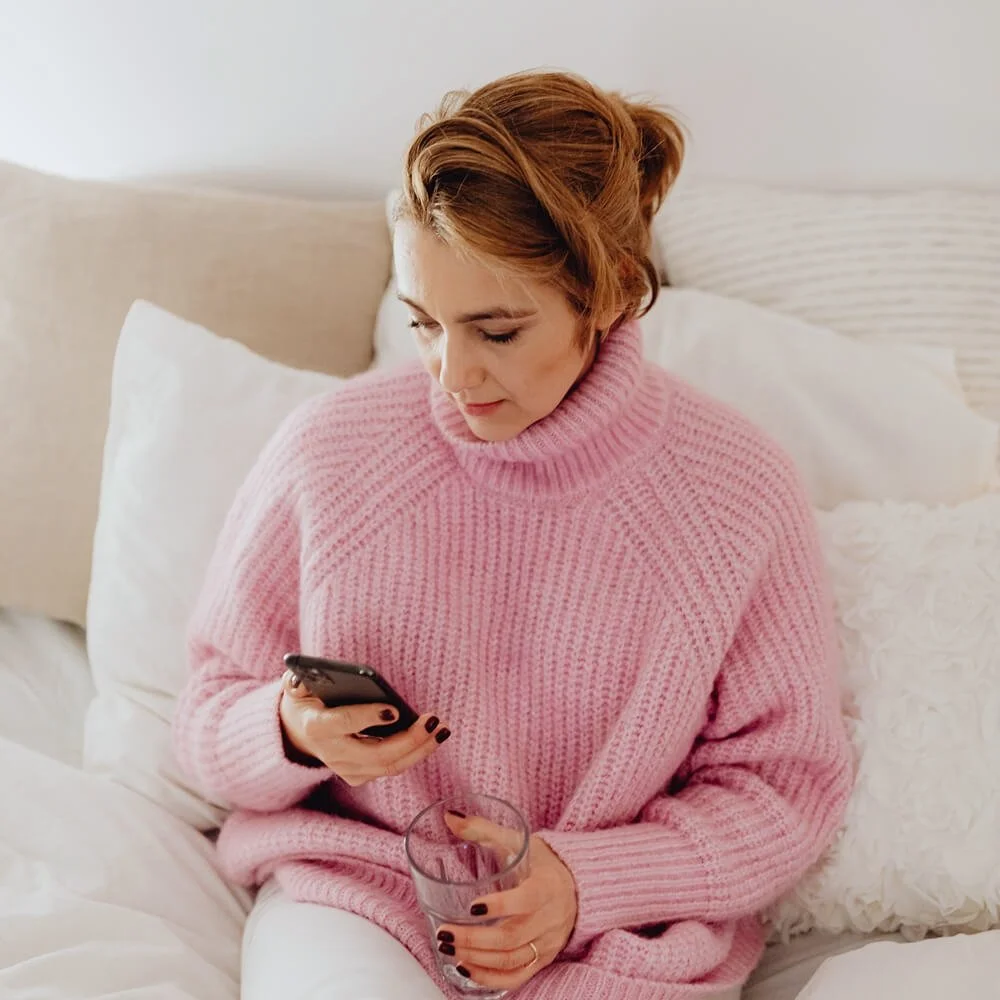 A woman with short reddish hair sitting on a bed, wearing a pink knitted turtleneck sweater, holding a glass of water in one hand and looking at her phone in the other.