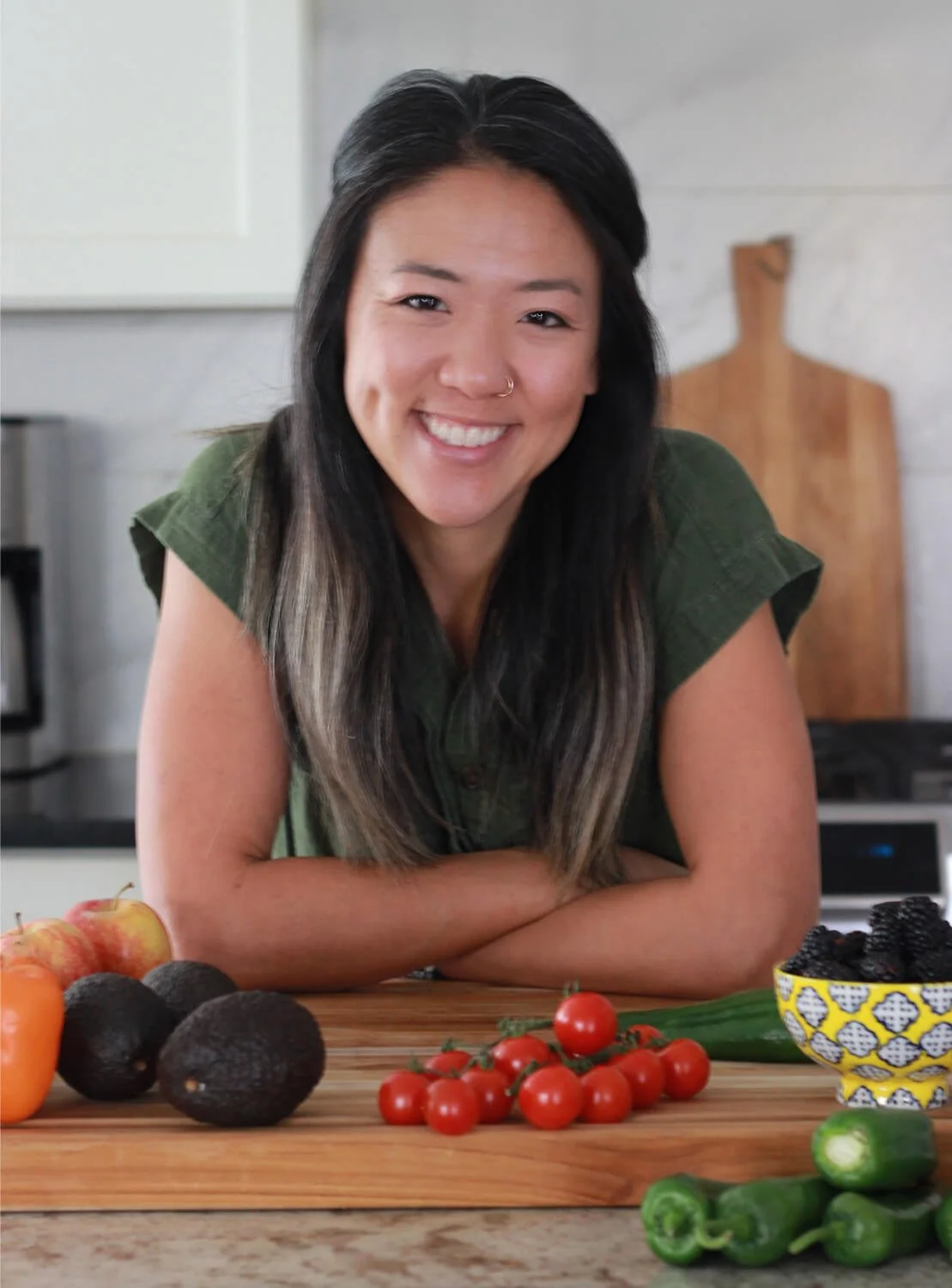 A woman with long black hair and a nose ring smiling at the camera, leaning on a wooden kitchen counter with fresh produce including tomatoes, blackberries, avocados, and cucumbers in front of her.