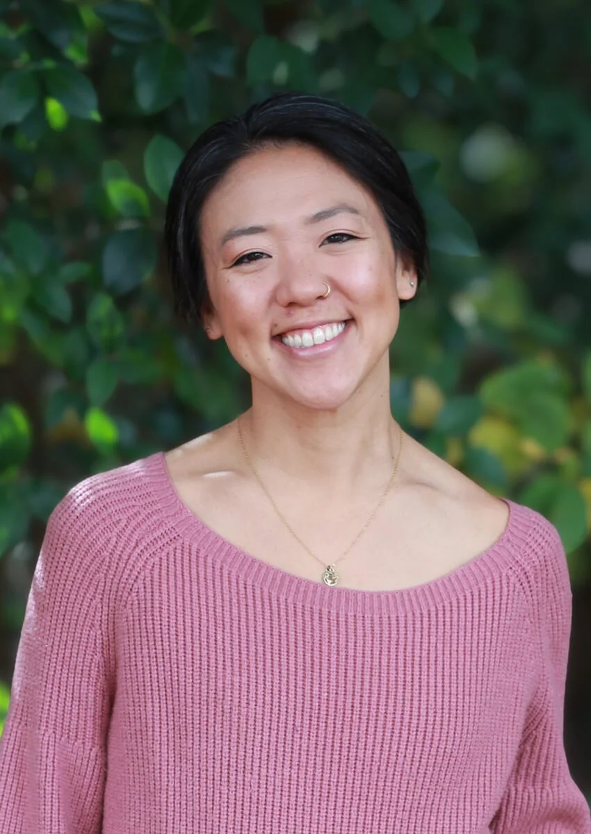 A smiling woman with short dark hair, wearing a pink knit sweater, a gold necklace, and a nose ring, standing outdoors in front of green foliage.