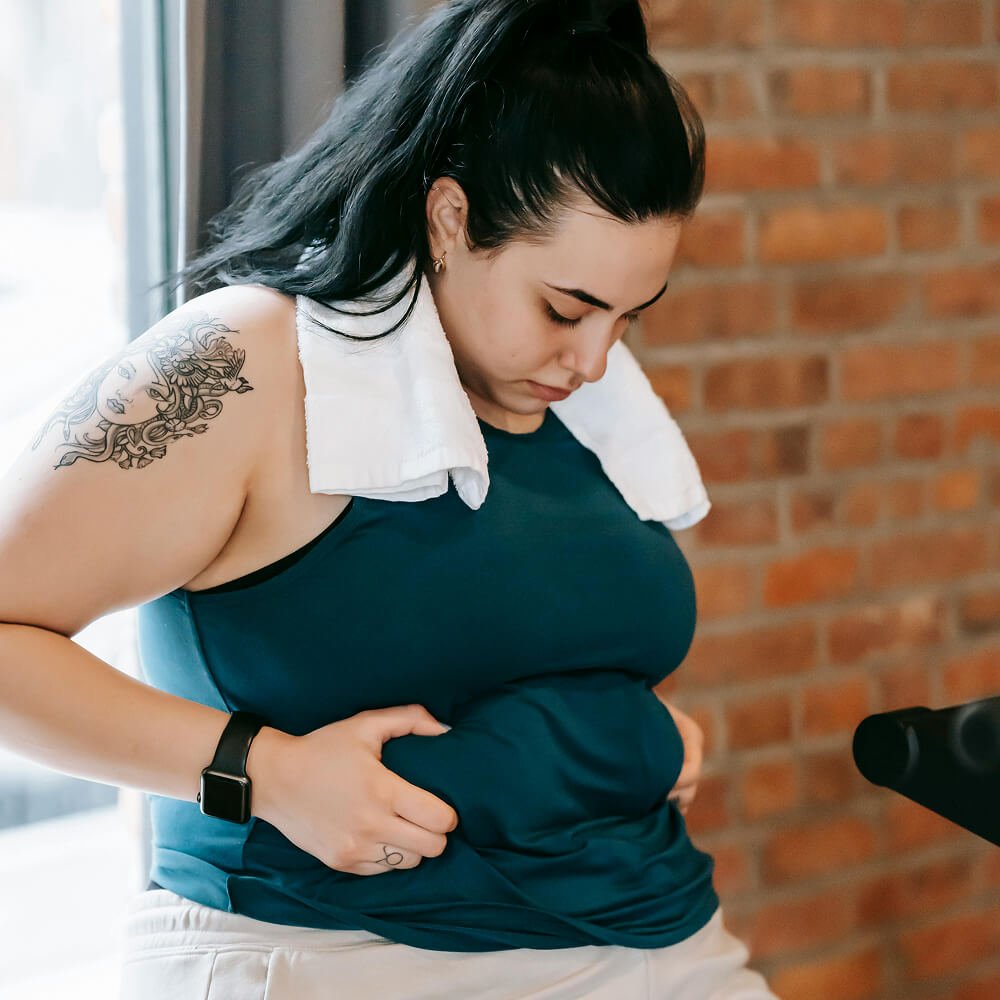 Woman in a blue tank top sitting, with a towel draped around her neck, looking down, with a brick wall in the background.