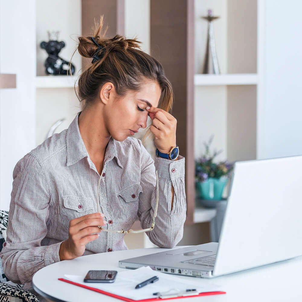 Woman sitting at a desk with a laptop, holding her forehead with one hand, looking stressed, with glasses, smartphone, and office supplies on the desk.