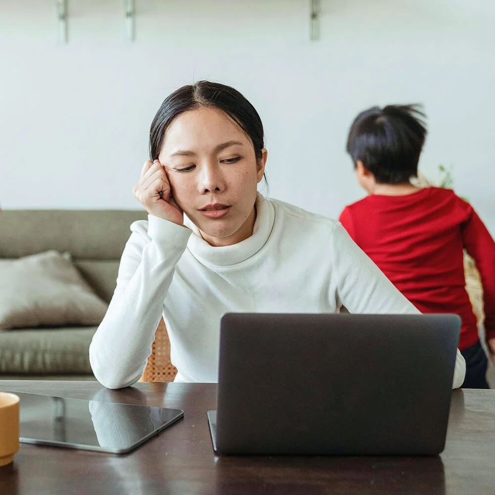 A woman in a white turtleneck sitting at a table, looking at a laptop with a thoughtful expression, and a boy in a red shirt in the background facing away from the camera.