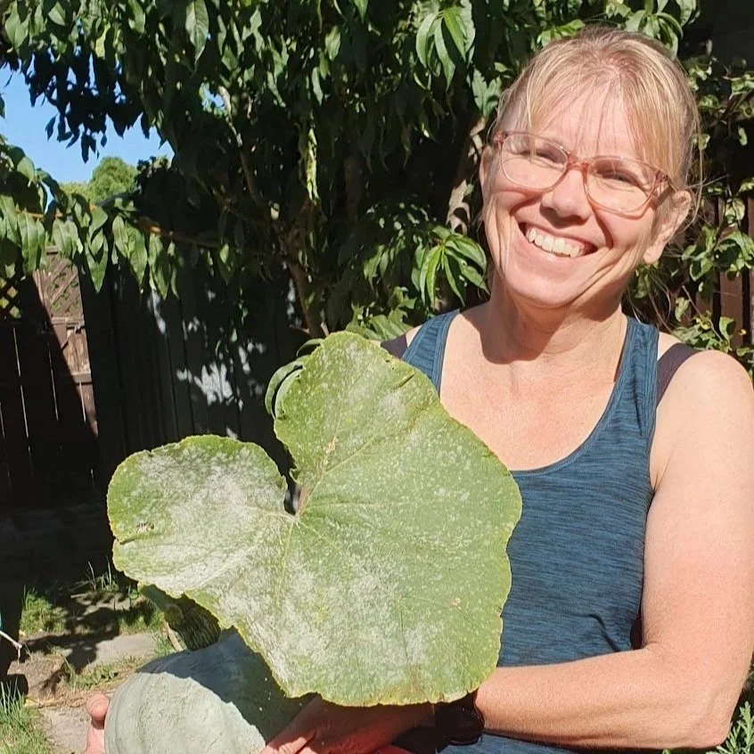 A woman with glasses and blonde hair smiling outdoors, holding a large green lotus leaf.