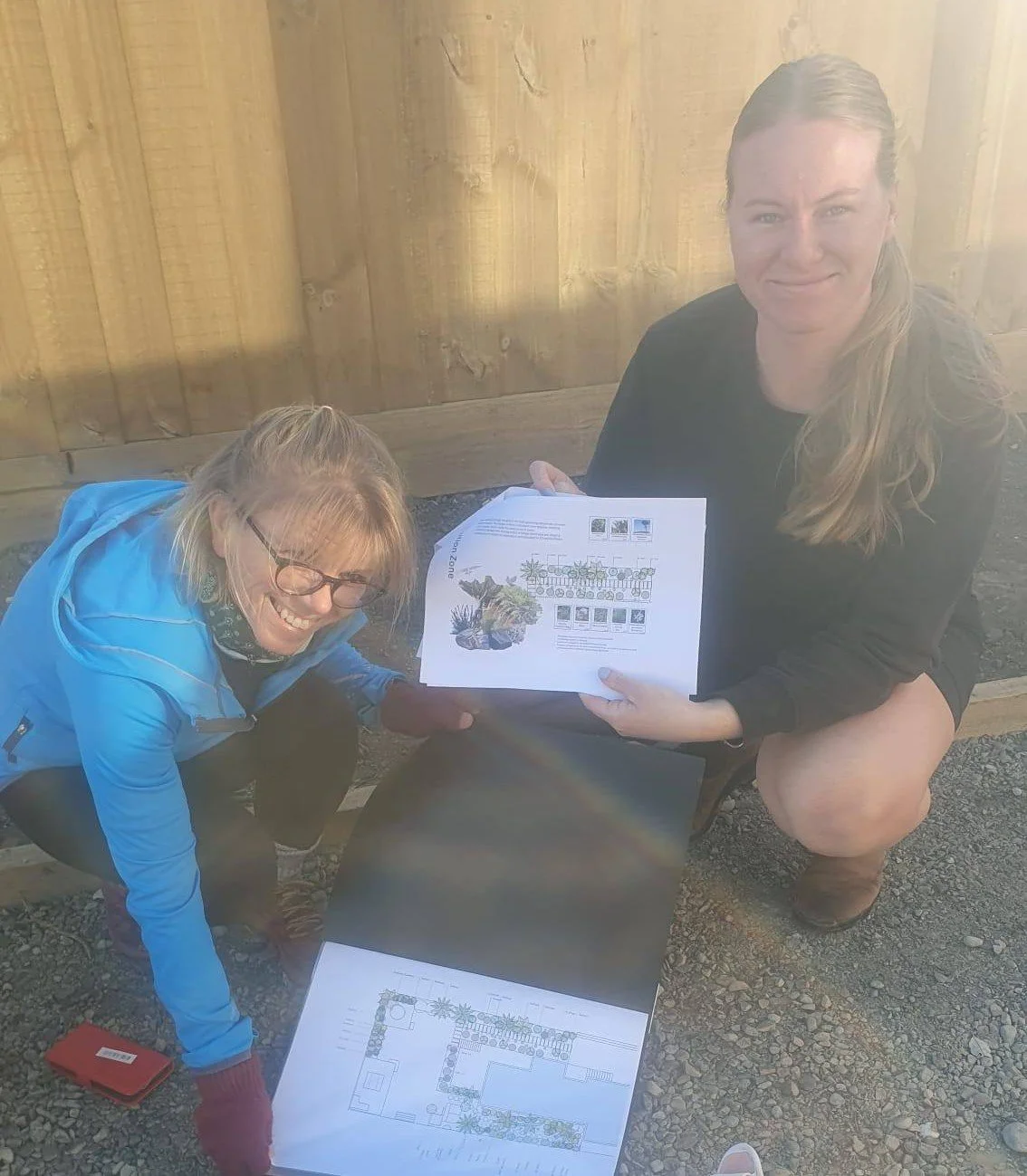 Two women outdoors near a wooden fence, holding and revealing large landscape or garden plans, smiling at the camera. Alannah Bee Garden Landscape Design Christchurch