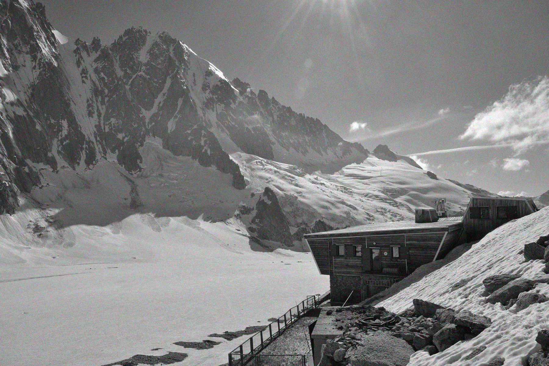 Cabane en bois dans un paysage de montagnes enneigées avec glacier, sous un ciel en partie nuageux et un soleil éclatant.
