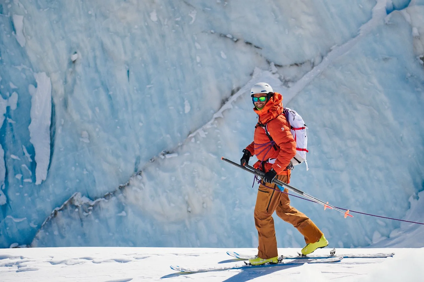 Une personne en ski avec un casque, lunettes de soleil et une veste orange, avançant dans un paysage glaciaire.