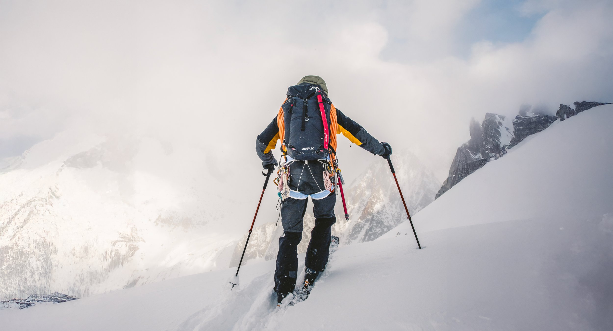 Personne faisant de la randonnée en montagne enneigée avec un sac à dos et des bâtons dans un paysage de montagnes couvertes de neige et de nuages.