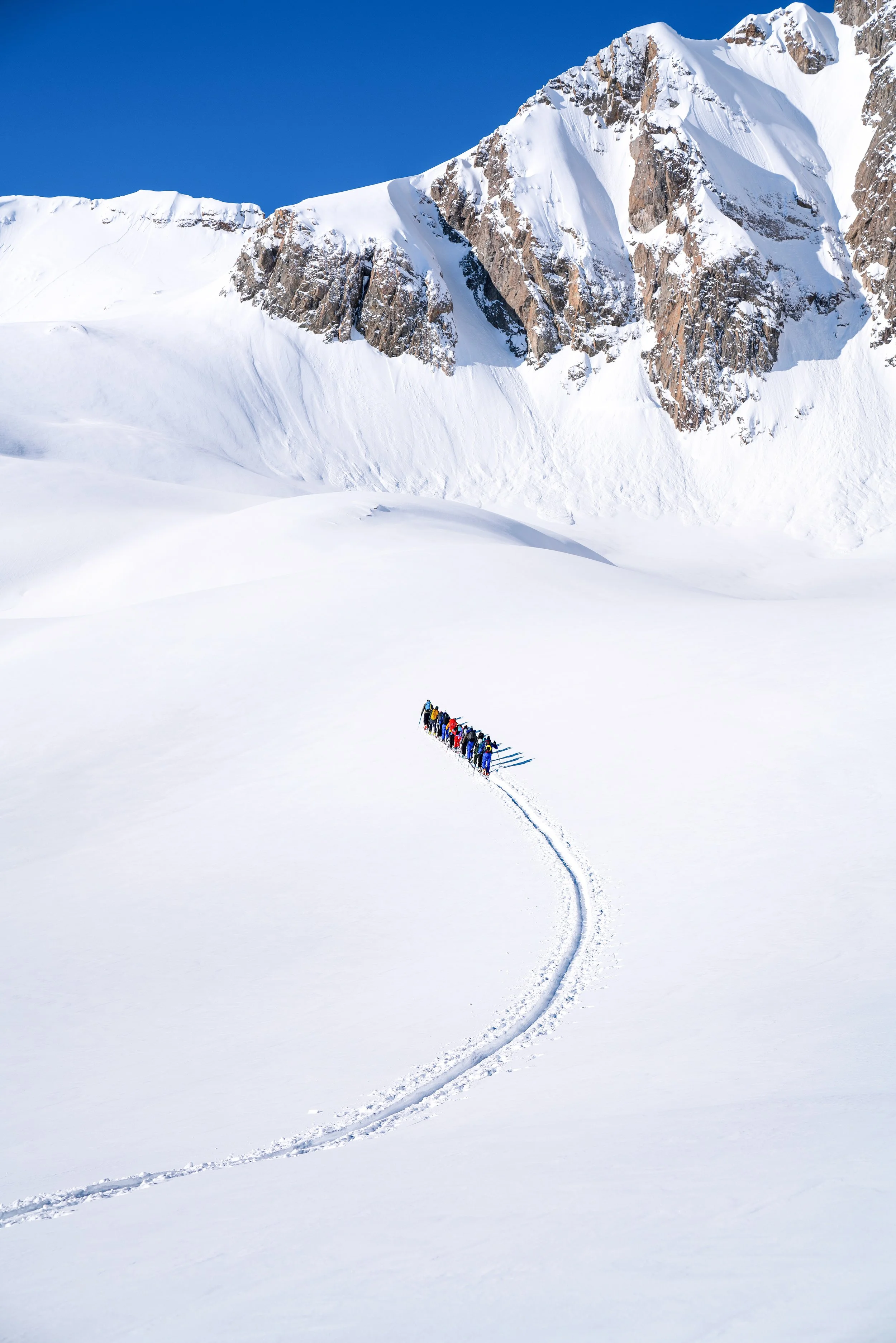 Groupe de personnes faisant une traversée dans la neige au pied de montagnes enneigées sous un ciel bleu clair