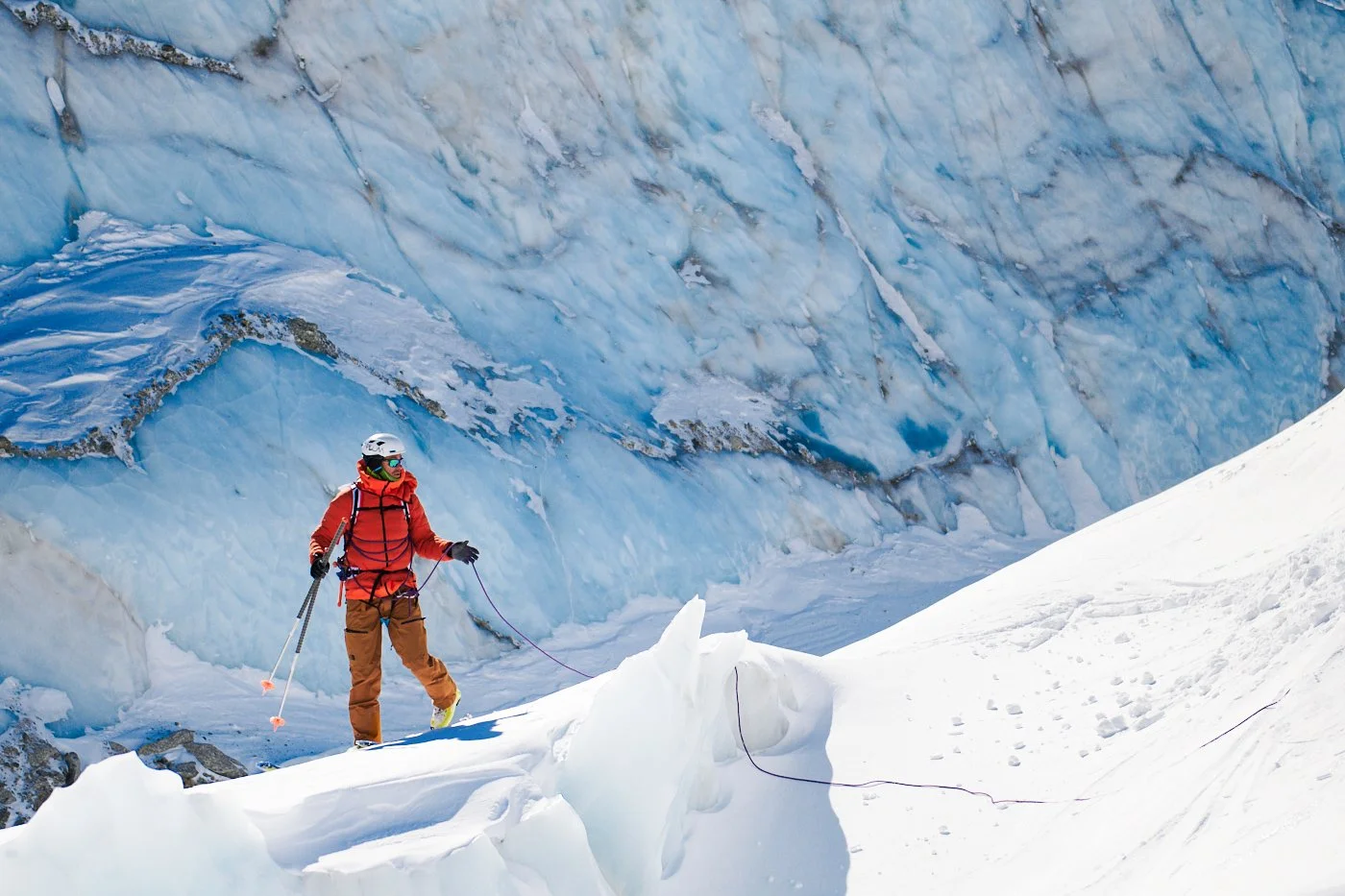 Un alpiniste en vêtements rouges et pantalons marron, portant un casque blanc et des lunettes, marche dans la neige près d'une calotte glacière bleue, tenant des bâtons de ski, dans un environnement glaciaire.