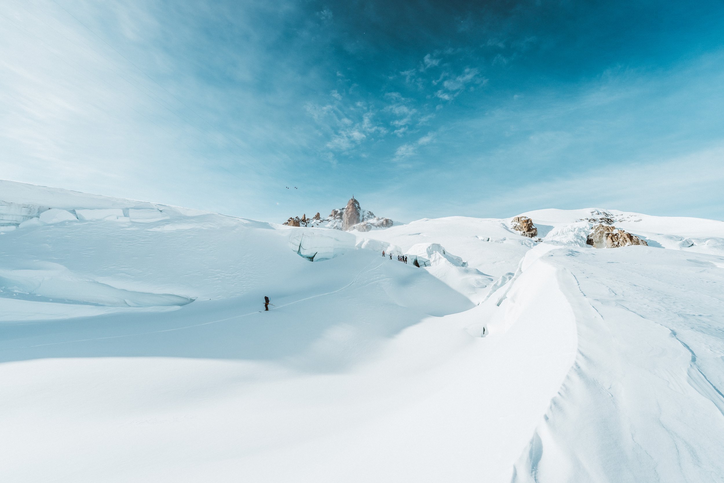 Paysage glacé avec des glaciers, des montagnes en arrière-plan et un ciel bleu clair, avec quelques personnes marchant dans la neige.
