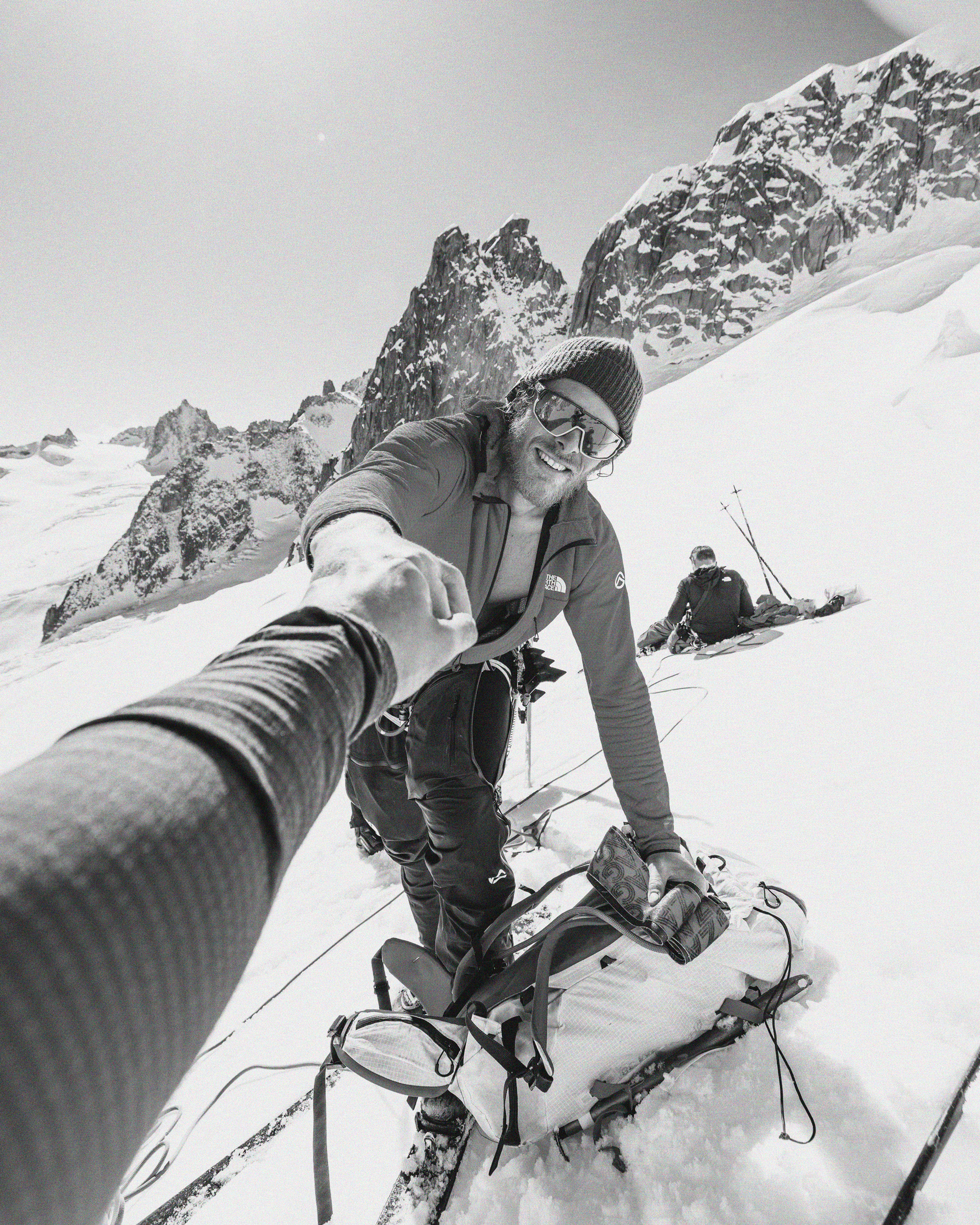 Deux alpinistes en haute montagne enneigée, avec des montagnes rocheuses en arrière-plan. Un homme en avant, portant des lunettes de ski et un manteau, tendant la main pour se faire aider, un autre homme repose sur le sol en arrière. Il y a des équipements de montagne autour, comme des skis, des bâtons et un sac à dos.