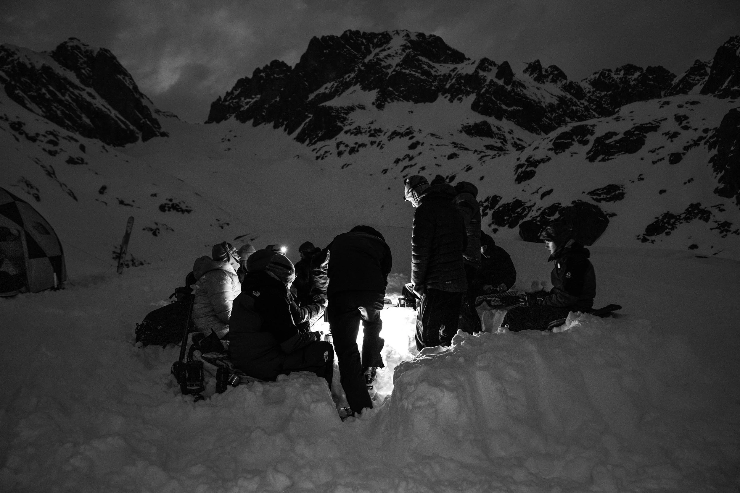 Groupe de personnes réunies autour d'une source de lumière dans un paysage enneigé de montagne, en noir et blanc, la nuit.