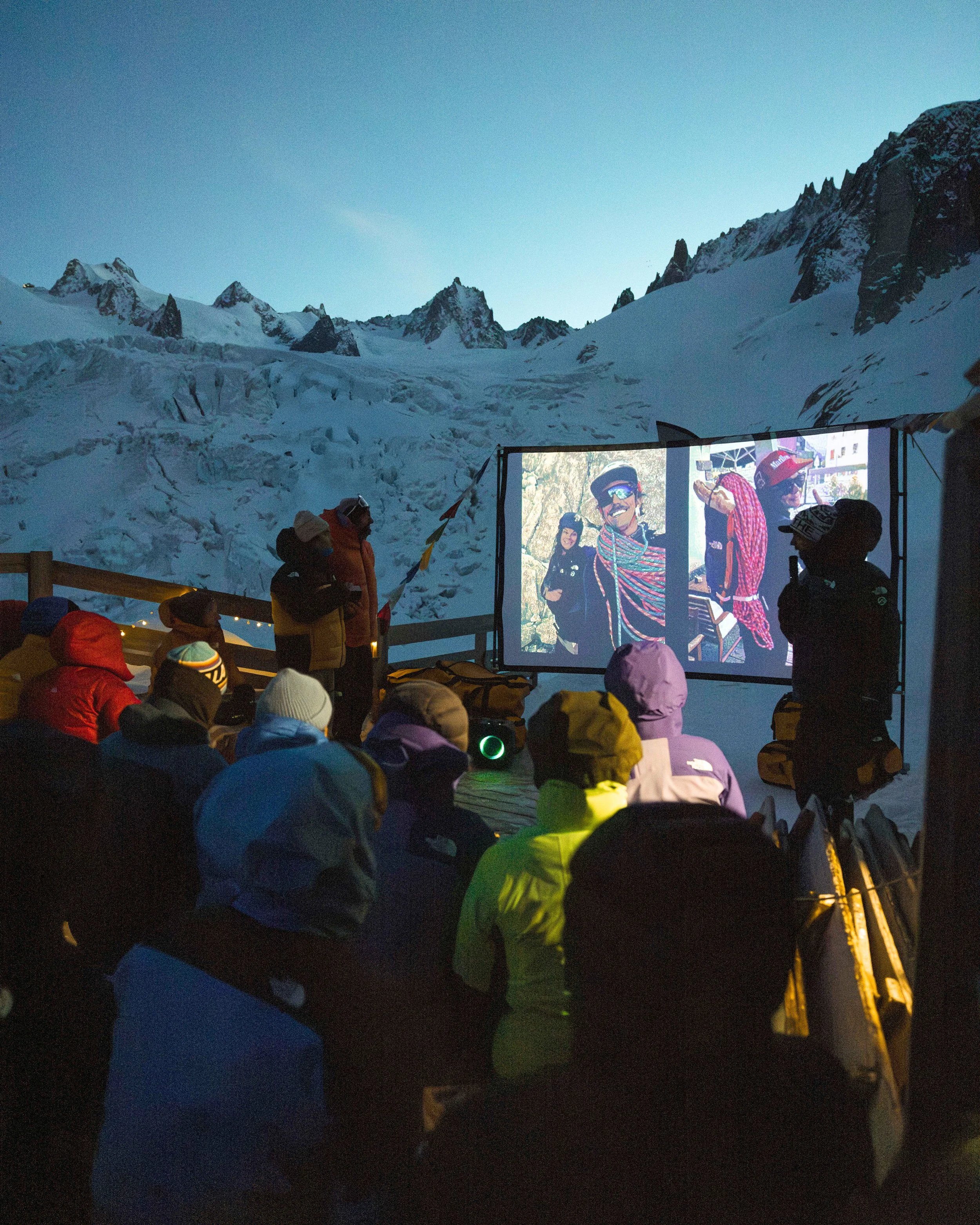 Groupe de personnes assises et regardant une projection en plein air, avec des montagnes enneigées en arrière-plan, lors d'une soirée.