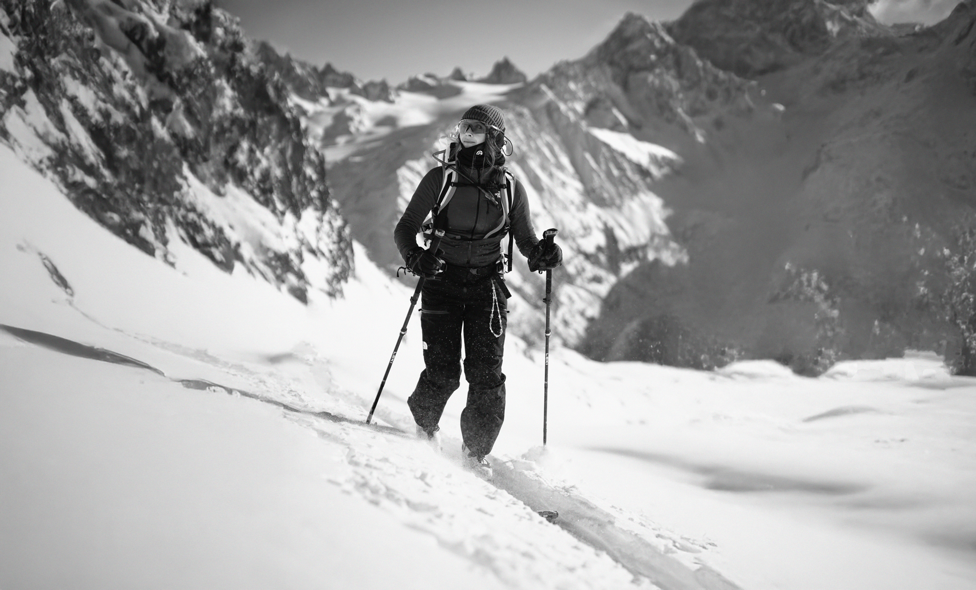 Une femme en vêtements de ski avec des bâtons dans la neige en montagne, portant un sac à dos, un bonnet et des lunettes, sous un ciel clair.