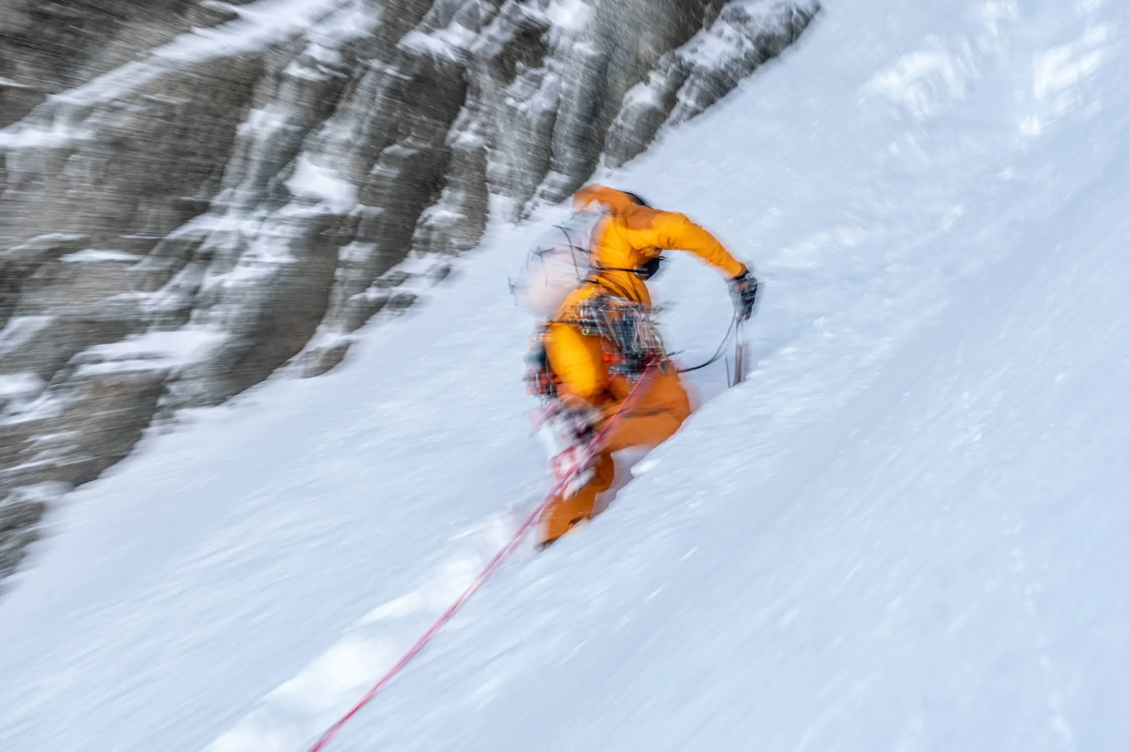 Un alpiniste en tenue orange grimpe une pente enneigée escarpée, avec des rochers visibles sur le côté gauche, lors d'une expédition en montagne ou en pleine nature enneigée.