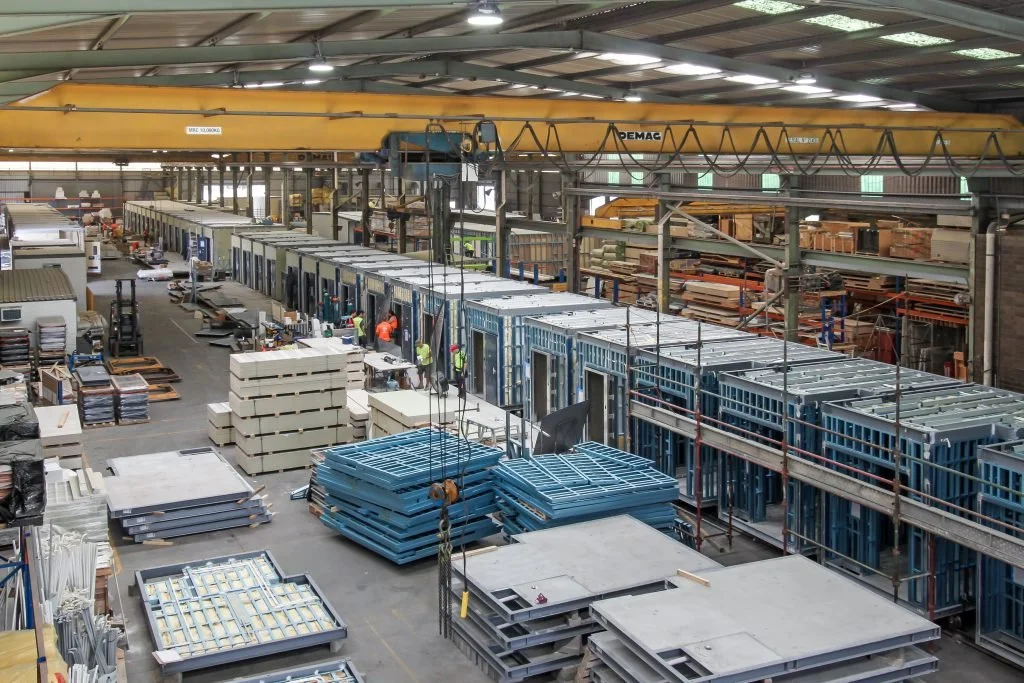 Inside a large industrial warehouse with construction workers assembling modular structures, with various building materials and equipment stored around.