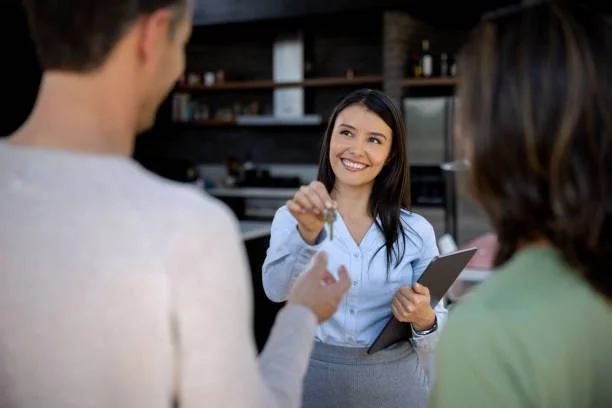 A woman smiling and handing a key to a man in a light-colored sweater, with another woman in a green top watching, in a modern kitchen setting.