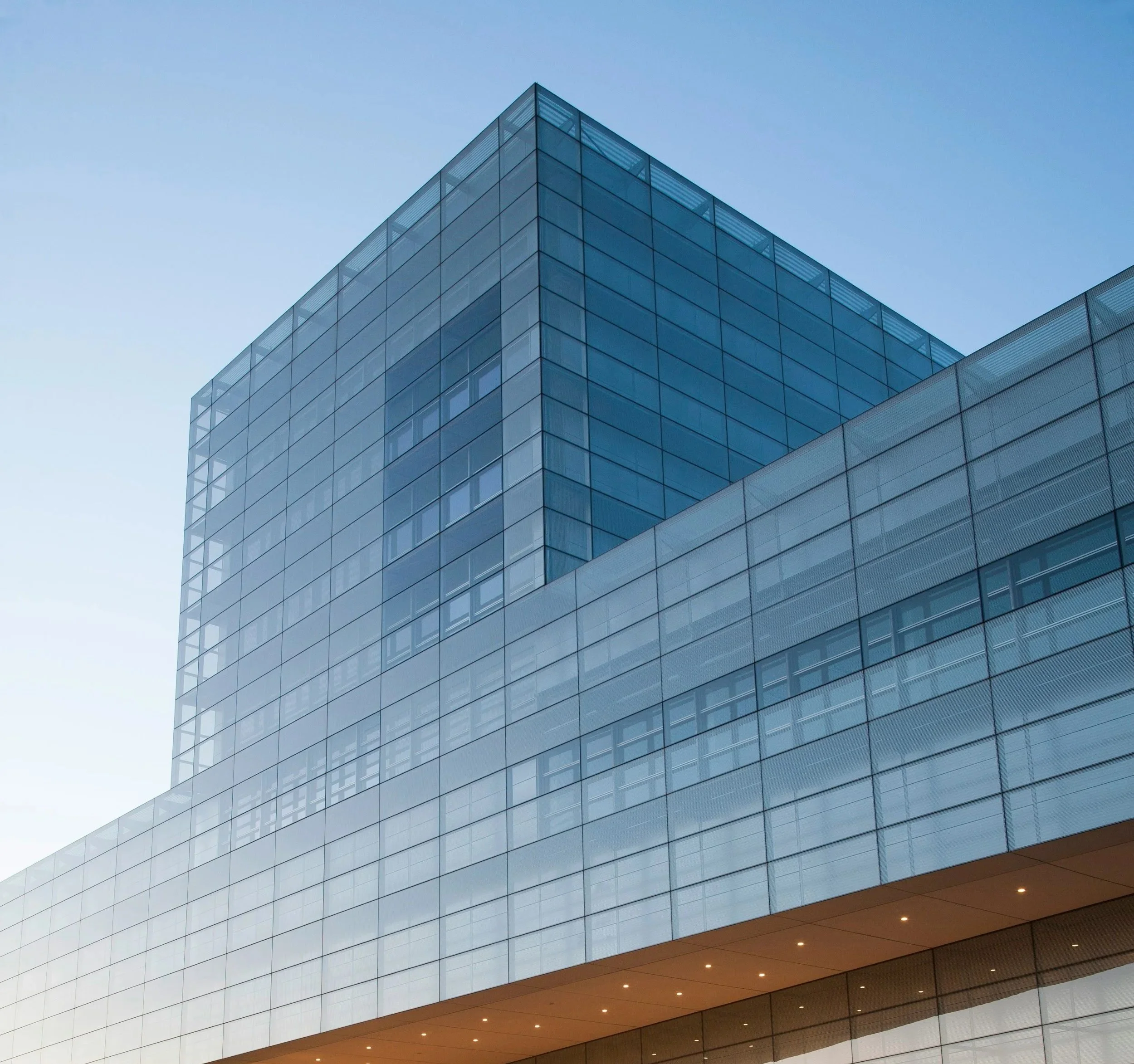 Modern glass building with multiple floors, reflecting the blue sky, with bright lighting at the bottom.