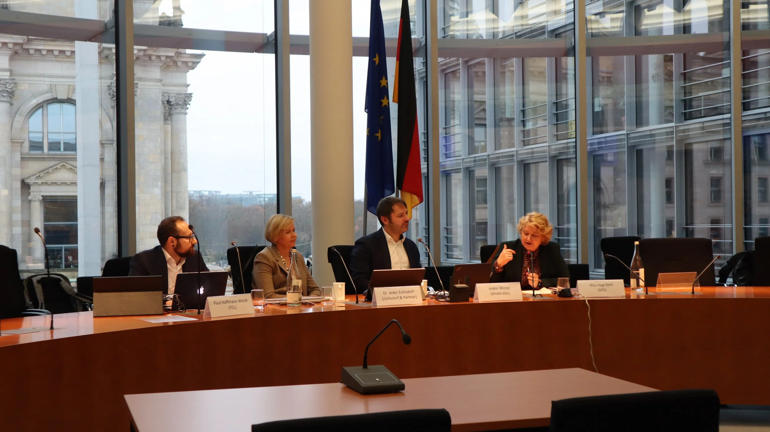 A panel of five people seated at a long conference table in a modern room with large glass windows and flags behind them. The panel includes four men and one woman, with name plaques and microphones in front of them. The room has an urban background with classical architecture visible through the windows.