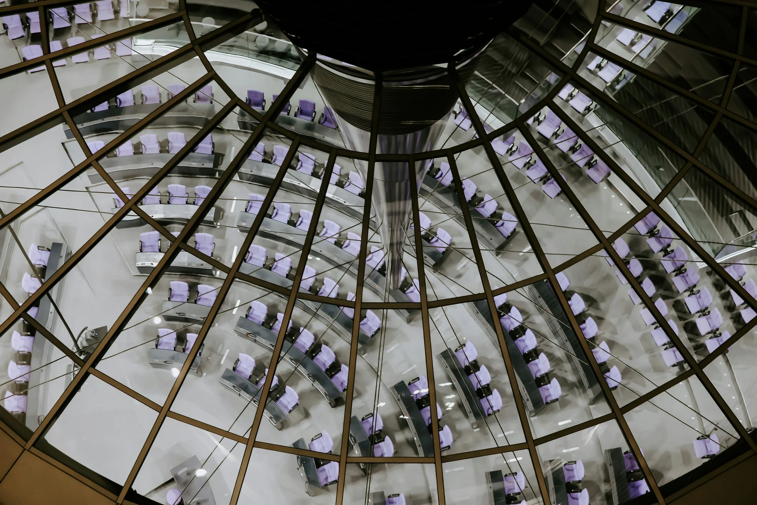 View looking down at an empty modern office space through glass panels, showing rows of purple chairs and workstations arranged in a circular pattern around a central column.