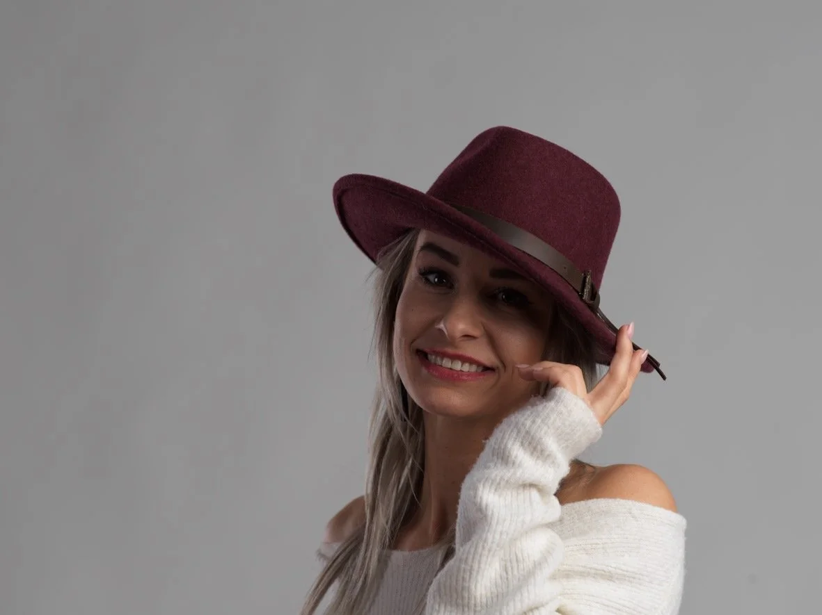 Vrouw met lang haar, die een bordeauxrode hoed draagt en een witte off-shoulder trui, glimlacht naar de camera.