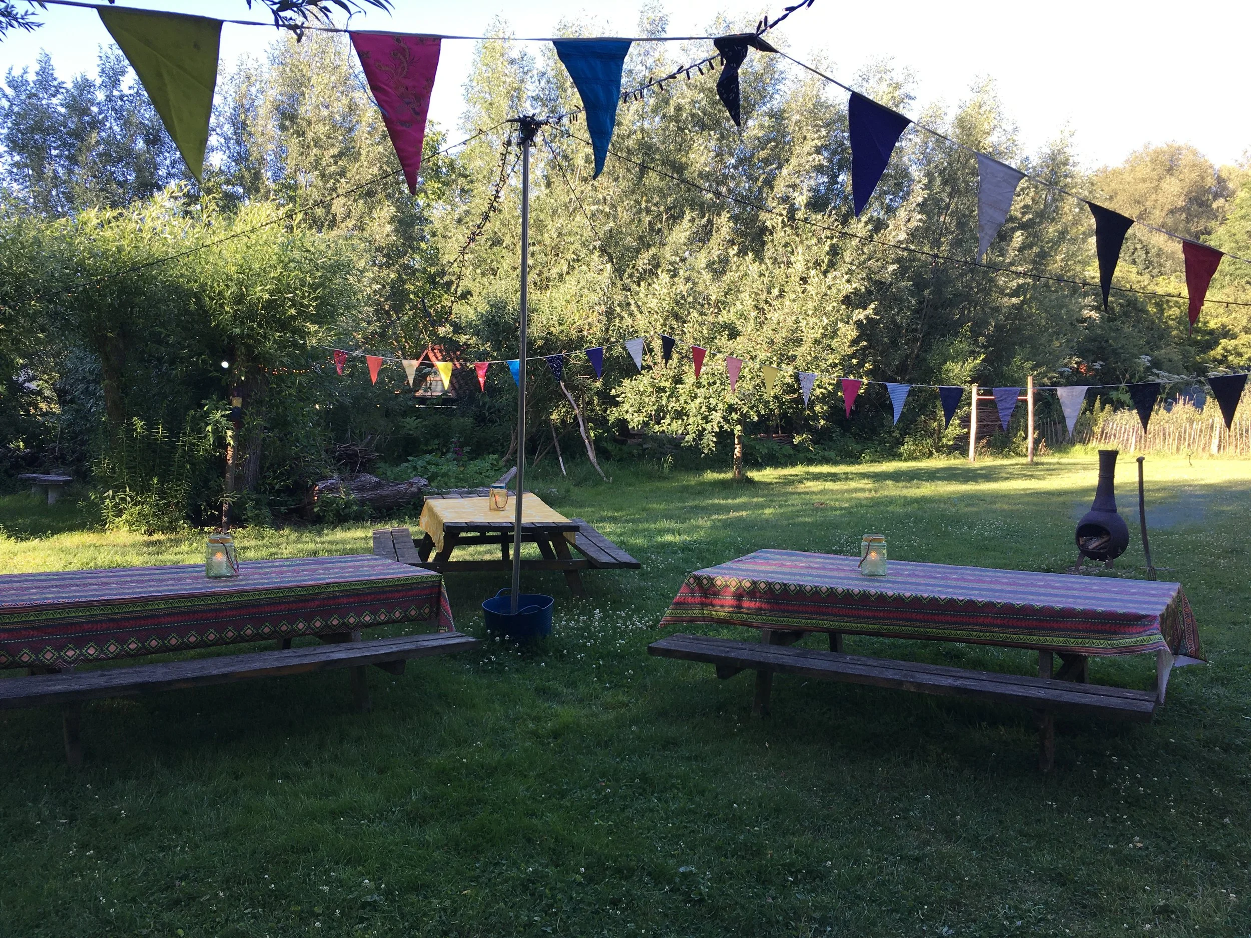 Buitentafel met kleurrijke tafelkleedjes in een tuin, versierd met vlaggetjes en een tuinovenschouw op de achtergrond, rondom groene bomen en gras.