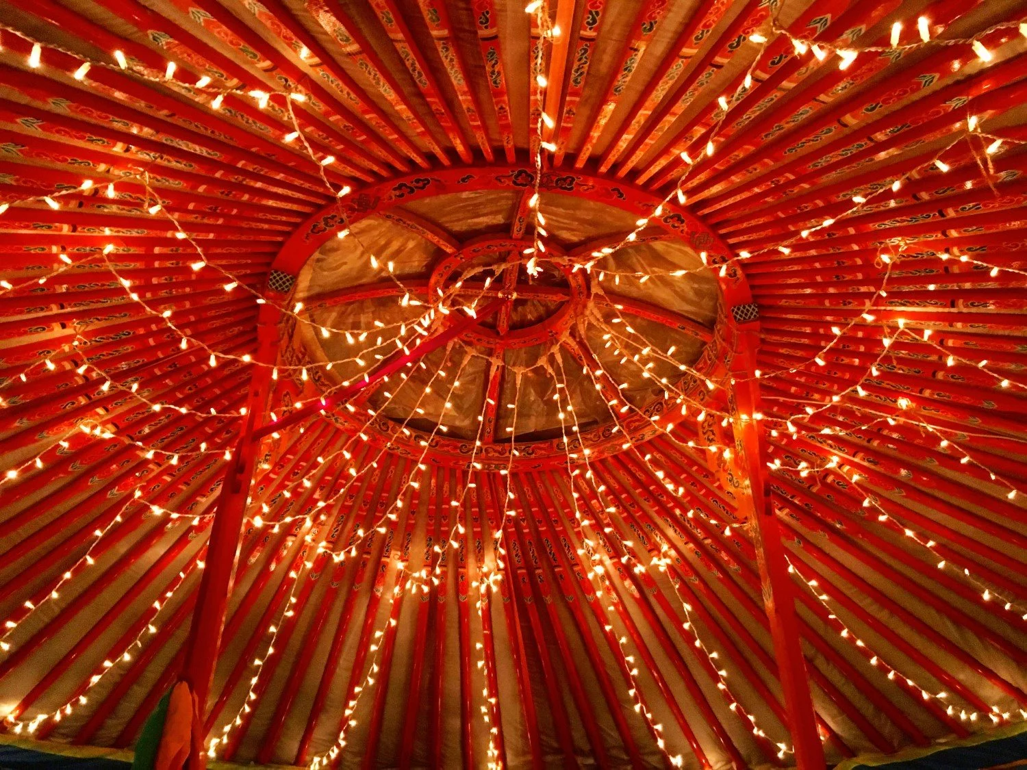 Het plafond van een traditionele yurt met rode stammen en sierlijke patronen, verlicht door kleine gezellige lichtjes.