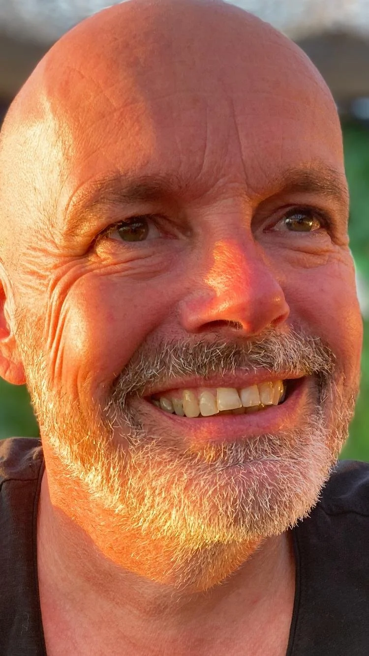 Close-up van een lachende man met een kale kop en een grijs-witte baard, shinende huid, ontspannen glimlach, in een natuurlijke omgeving bij zonsondergang.