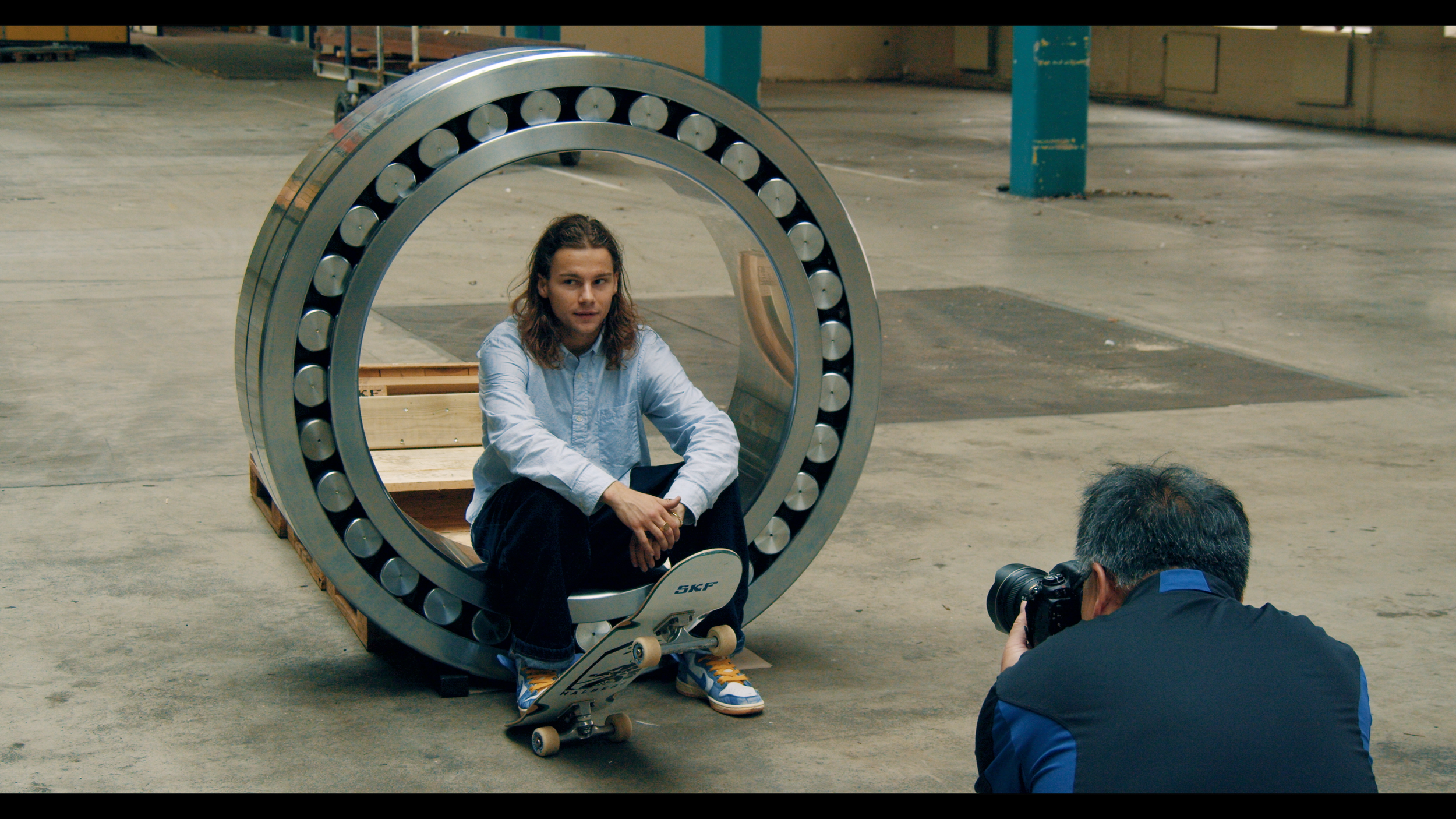 A man is taking a photo of a woman sitting inside a large metallic bearing in an industrial warehouse. The woman has long brown hair, is wearing a light blue shirt, and colorful sneakers.