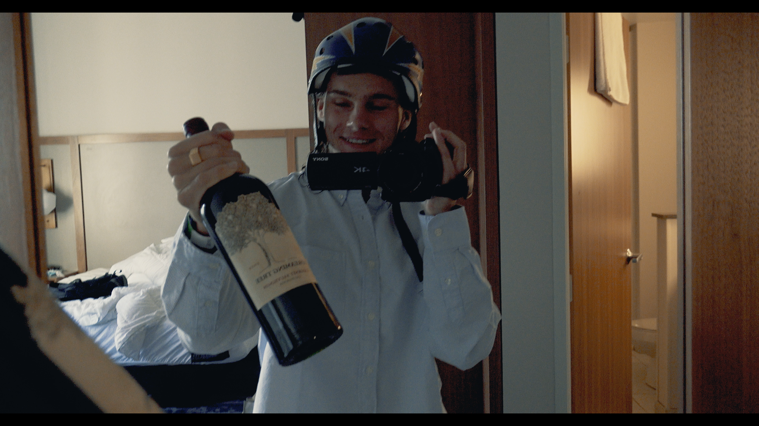 Person in a bicycle helmet holding a bottle of wine and taking a selfie in a mirror in a bedroom.