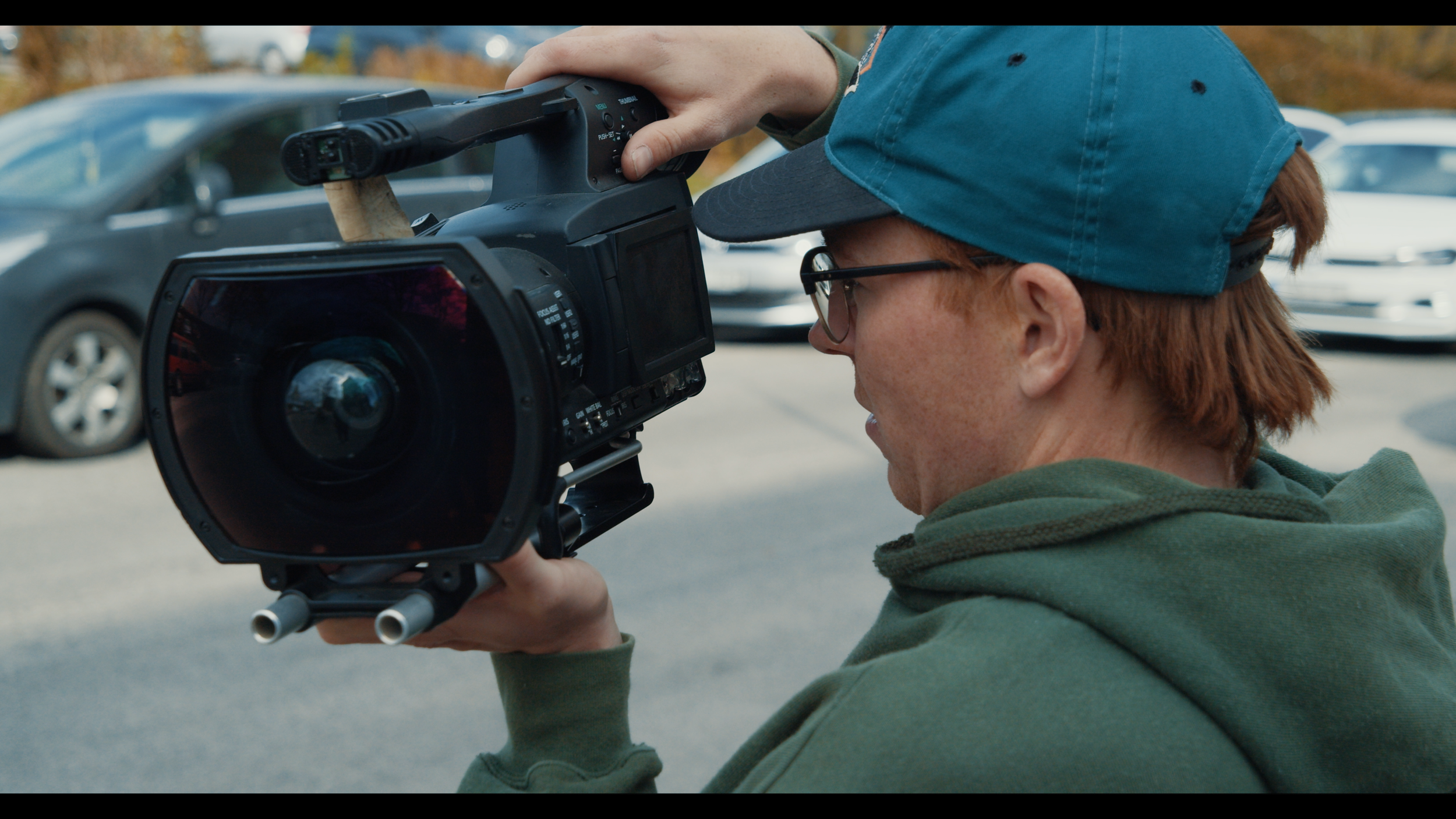 Person with glasses and a blue cap holding a professional camera on a tripod in an outdoor parking lot.