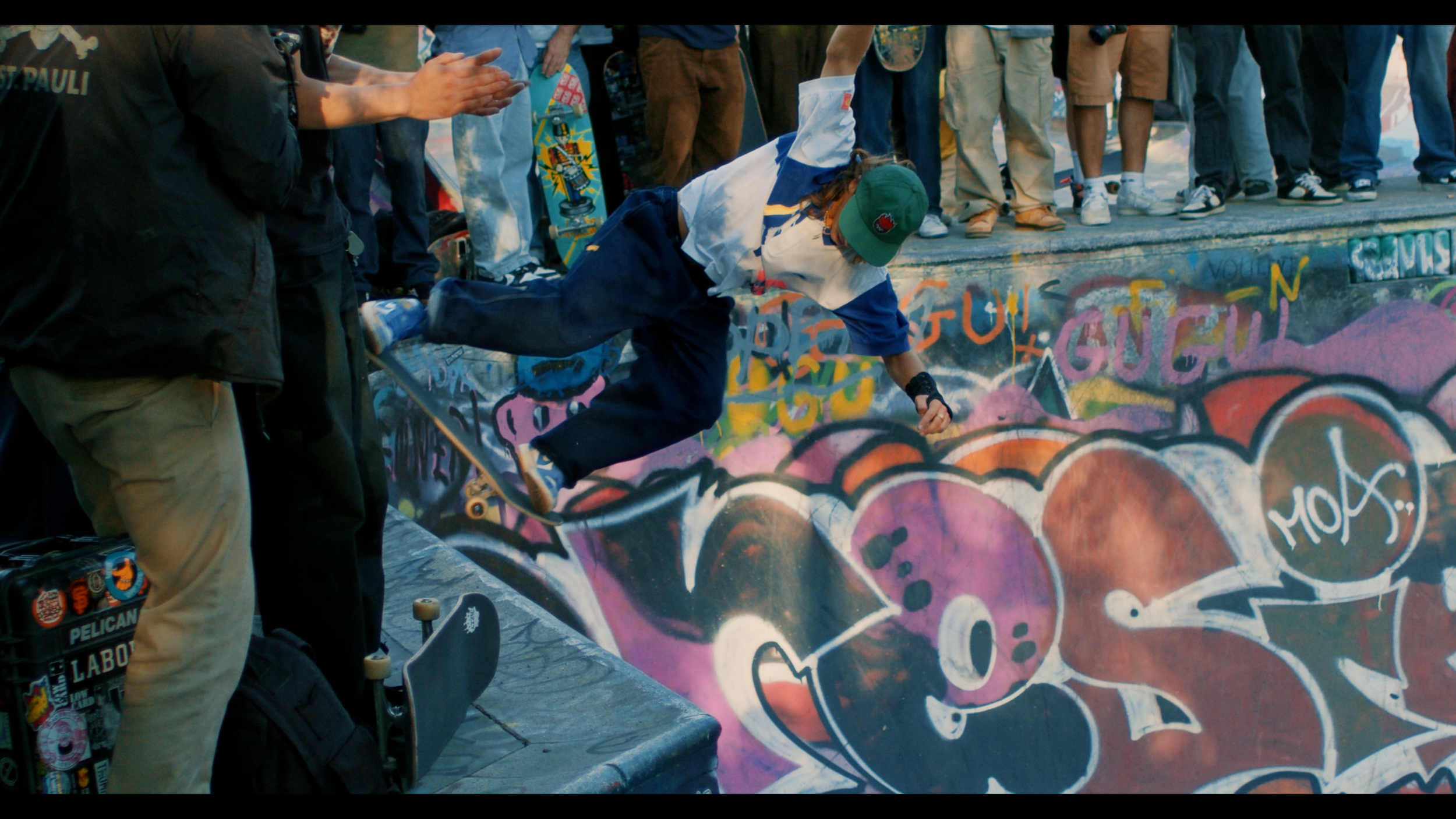 A skateboarder wearing a green cap and white jacket performing a trick in a graffiti-covered skate park while surrounded by onlookers.